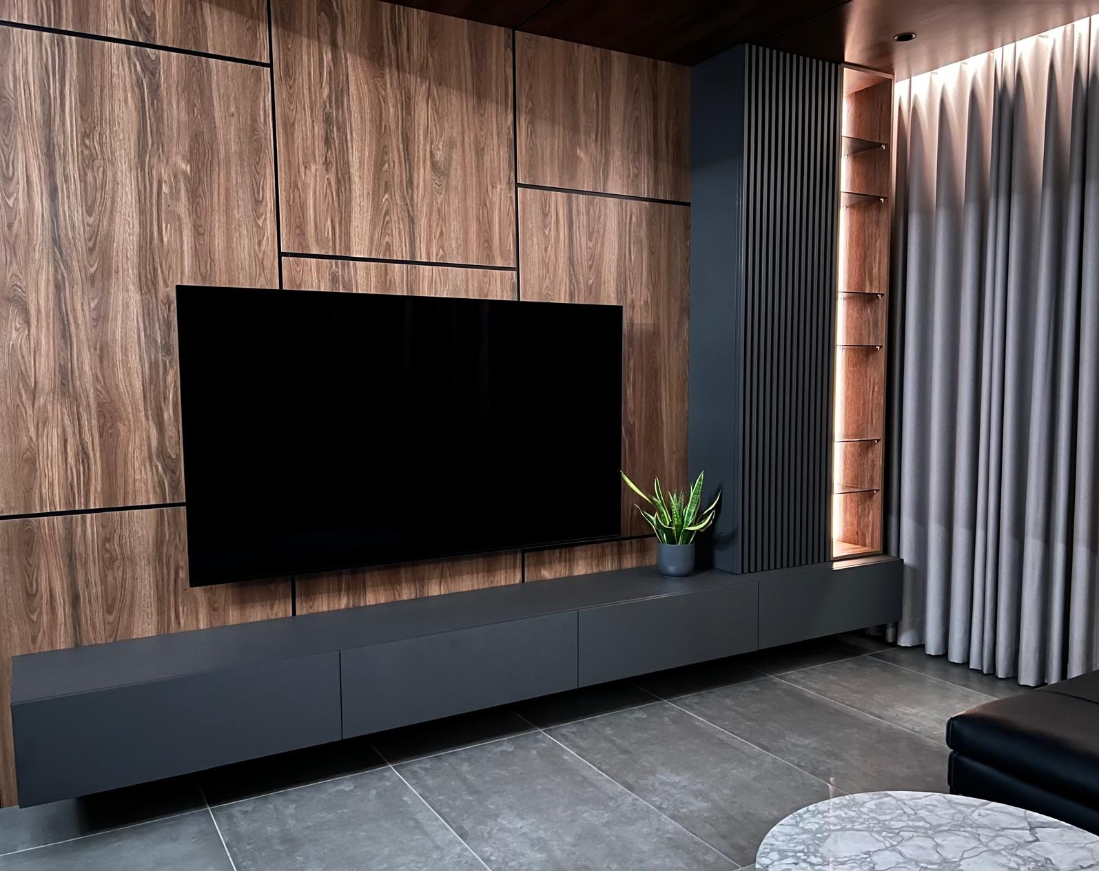 Nowoczesna ściana telewizyjna z drewnianymi panelami, czarnym telewizorem, szarą szafką RTV i podświetlaną półką, uzupełniona zieloną rośliną w doniczce.