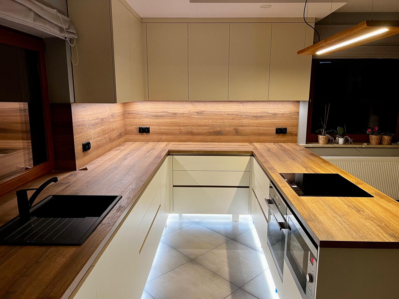 Nowoczesna kuchnia w kształcie litery U z beżowymi szafkami, drewnianym blatem i podświetleniem LED pod szafkami dolnymi, czarny zlew i płyta indukcyjna.