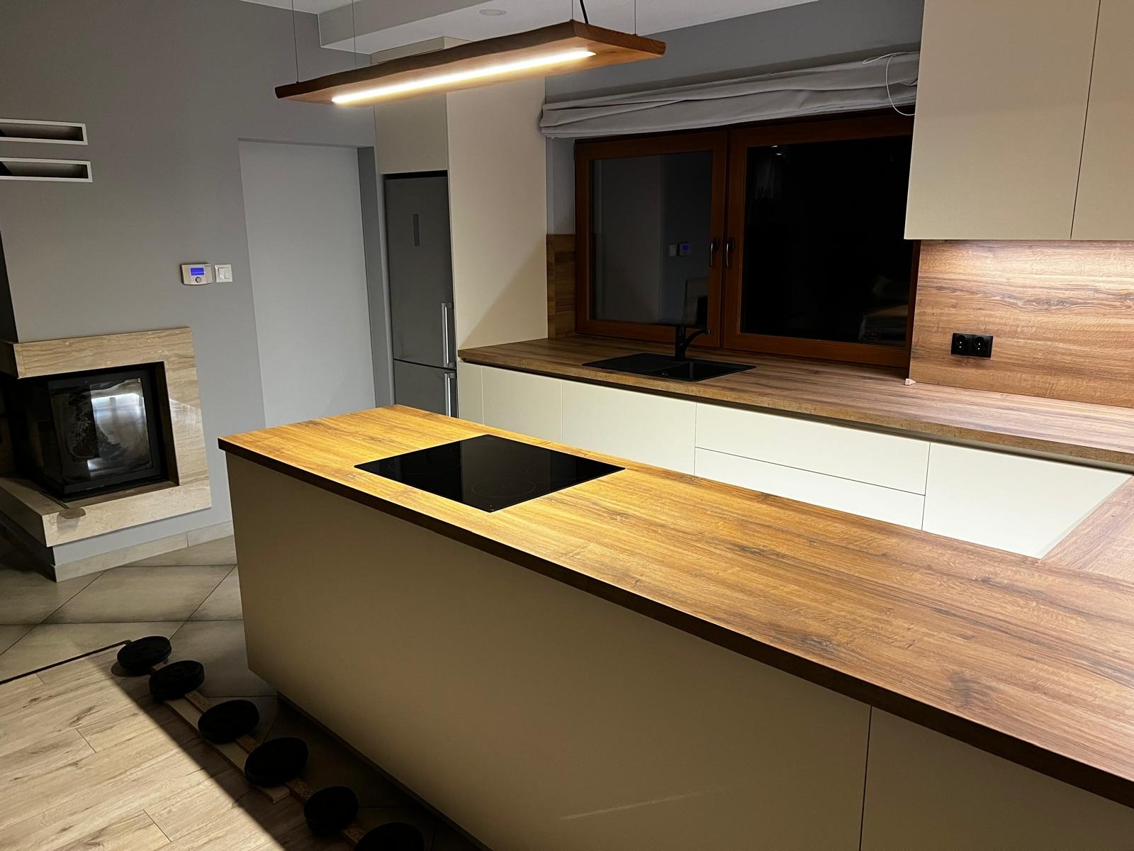 Nowoczesna kuchnia z wyspą, drewnianym blatem i płytą indukcyjną, minimalistyczne oświetlenie LED, widoczny kominek w tle.