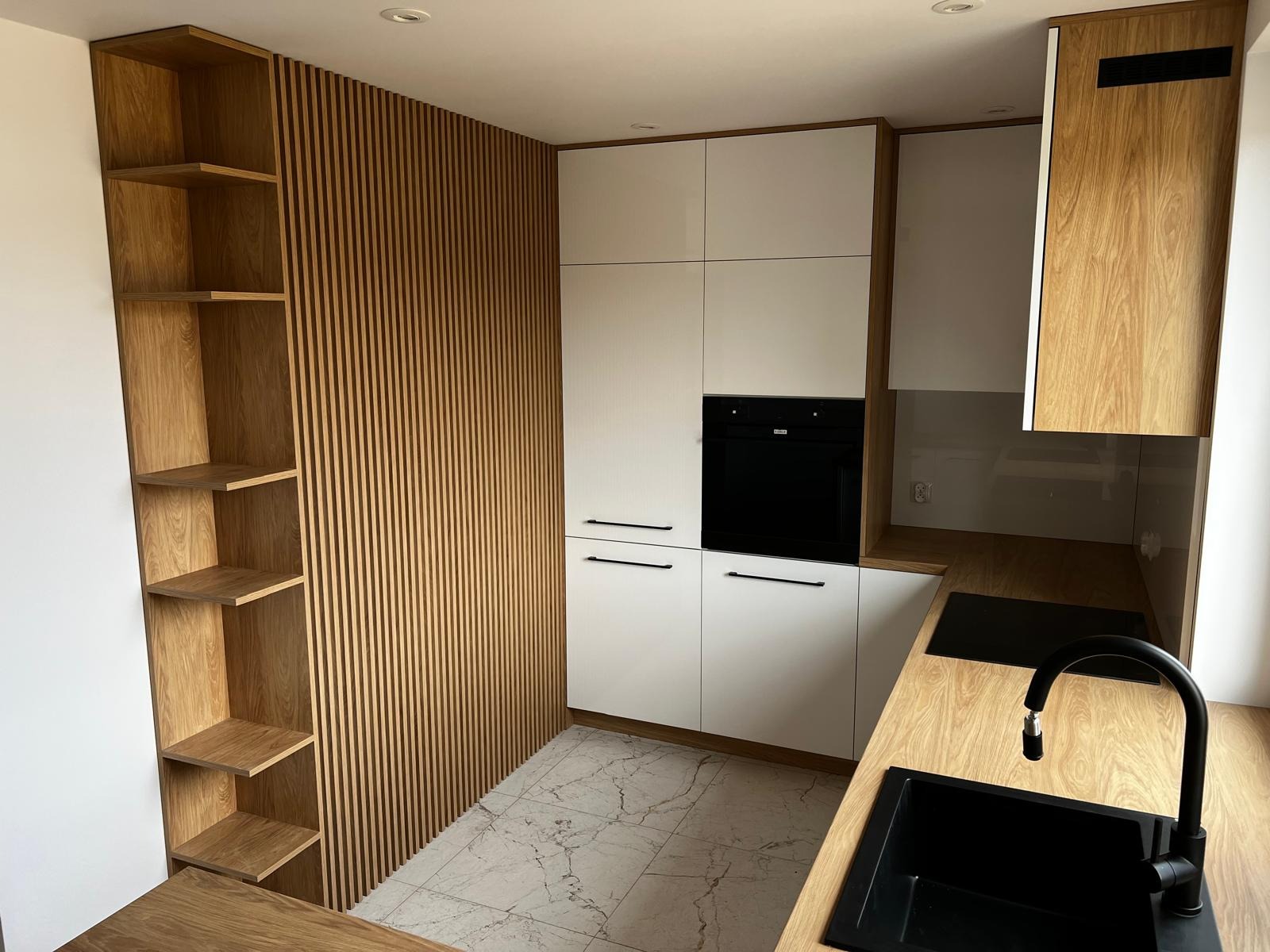 Nowoczesna kuchnia z białymi frontami szafek, czarnymi uchwytami i piekarnikiem w zabudowie, drewnianym blatem oraz ścianą z drewnianych listew i półkami.