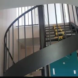 Metalowe schody kręcone wewnątrz budynku, zabezpieczone taśmą malarską na stopniach, widok z dołu na konstrukcję z pionowymi prętami.