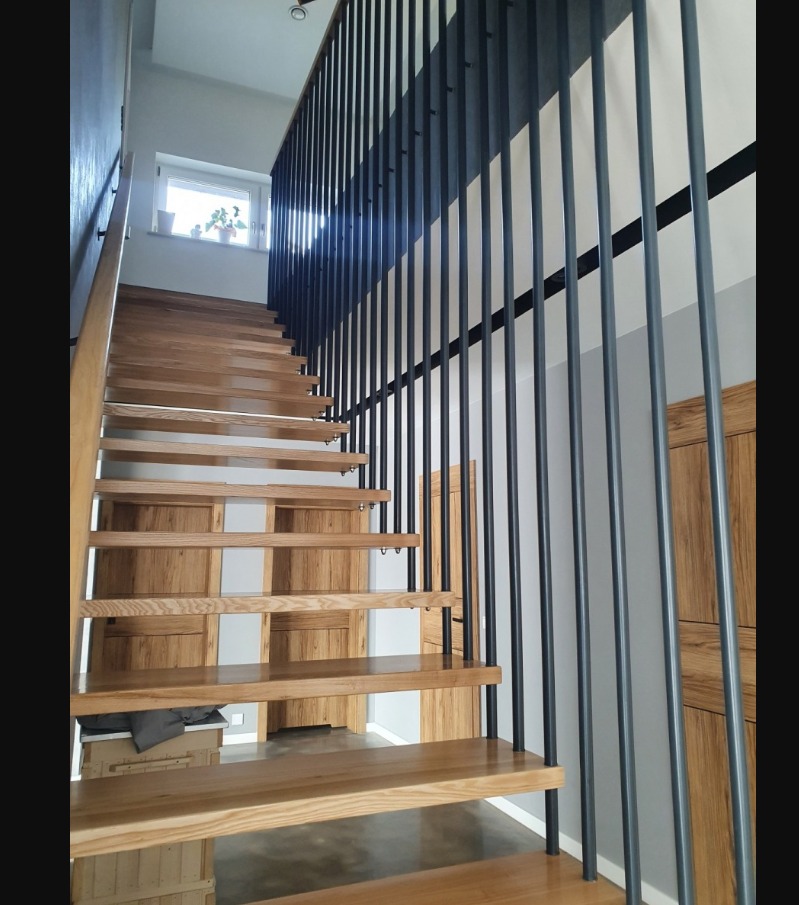 Drewniane schody z czarną, metalową balustradą z pionowych prętów, widoczne drzwi w tle, ujęcie od dołu schodów.