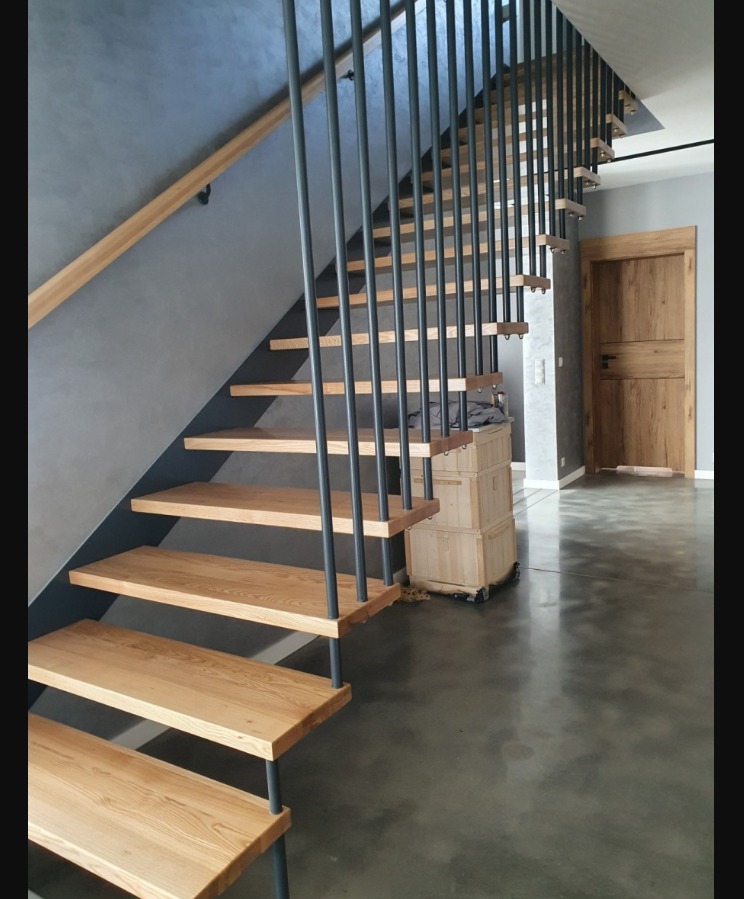 Nowoczesne schody z drewnianymi stopniami i czarnymi, metalowymi balustradami pionowymi, zamontowane w minimalistycznym wnętrzu z szarą ścianą i drewnianymi drzwiami.
