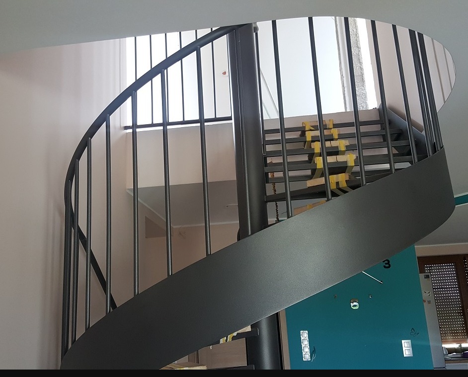 Metalowe schody kręcone wewnątrz budynku, zabezpieczone taśmą malarską na stopniach, widok z dołu na konstrukcję z pionowymi prętami.