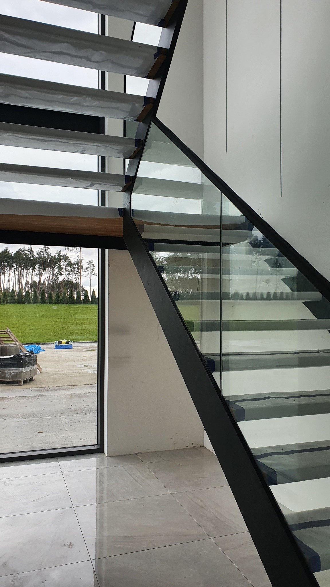 Wewnętrzne schody z metalową konstrukcją i szklaną balustradą, stopnie zabezpieczone folią ochronną, widok przez okno na zewnątrz.