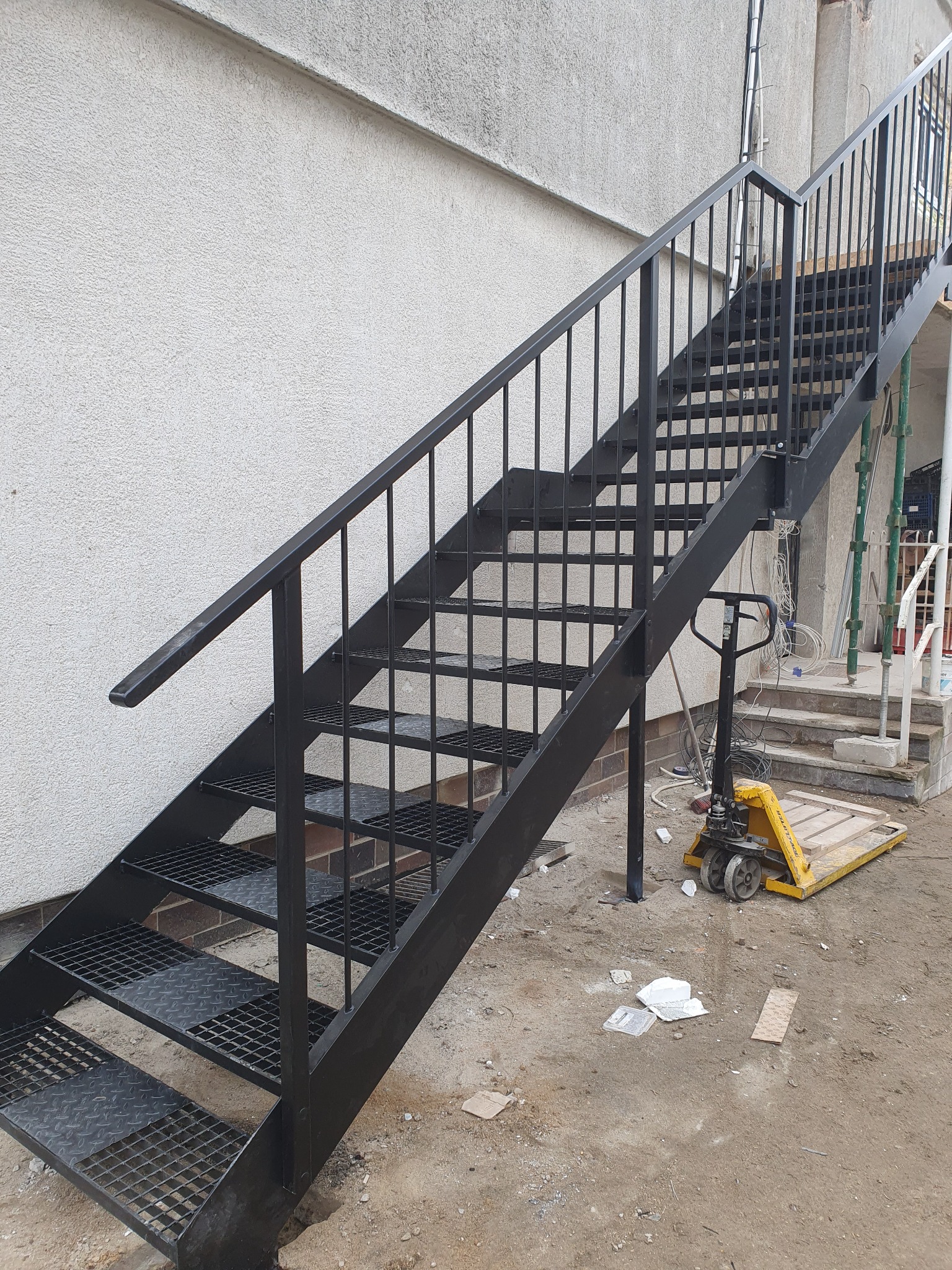 Zewnętrzne schody metalowe z czarną balustradą, prowadzące na wyższy poziom budynku, obok żółty wózek paletowy, widoczne ślady prac budowlanych.