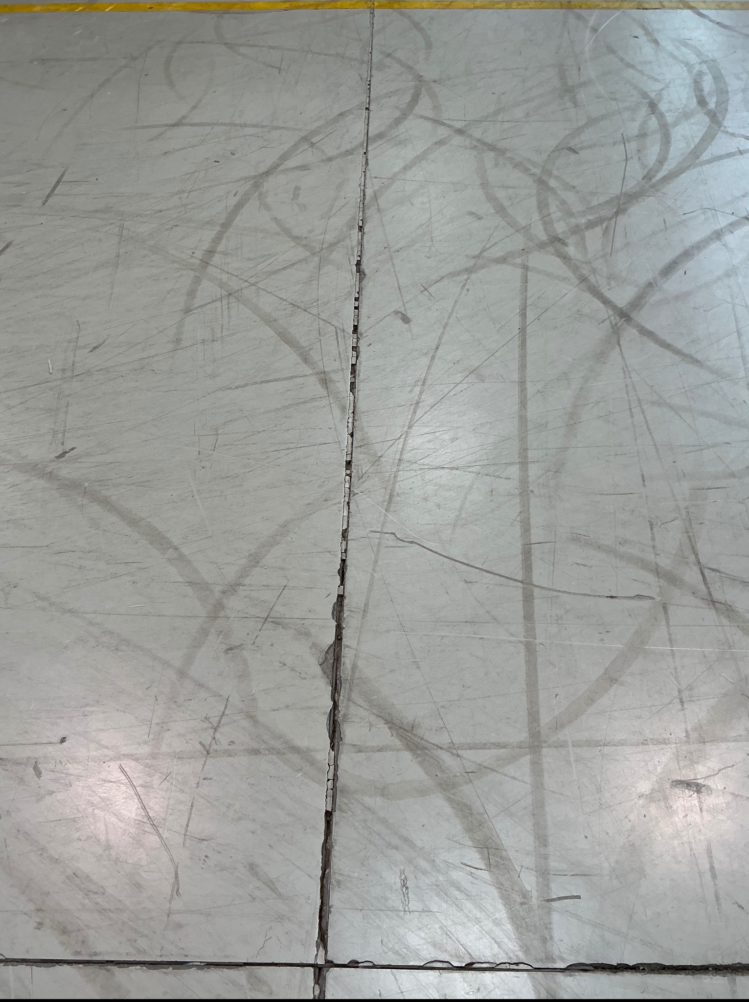 Szara, popękana podłoga z widocznymi śladami użytkowania i żółtą linią w górnej części kadru. Widoczne rysy i zabrudzenia.