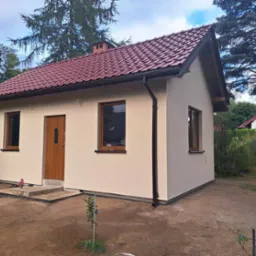Niewielki, ocieplony dom z beżową elewacją, brązowymi oknami i drzwiami oraz czerwoną dachówką, otoczony przygotowanym terenem pod ogród.