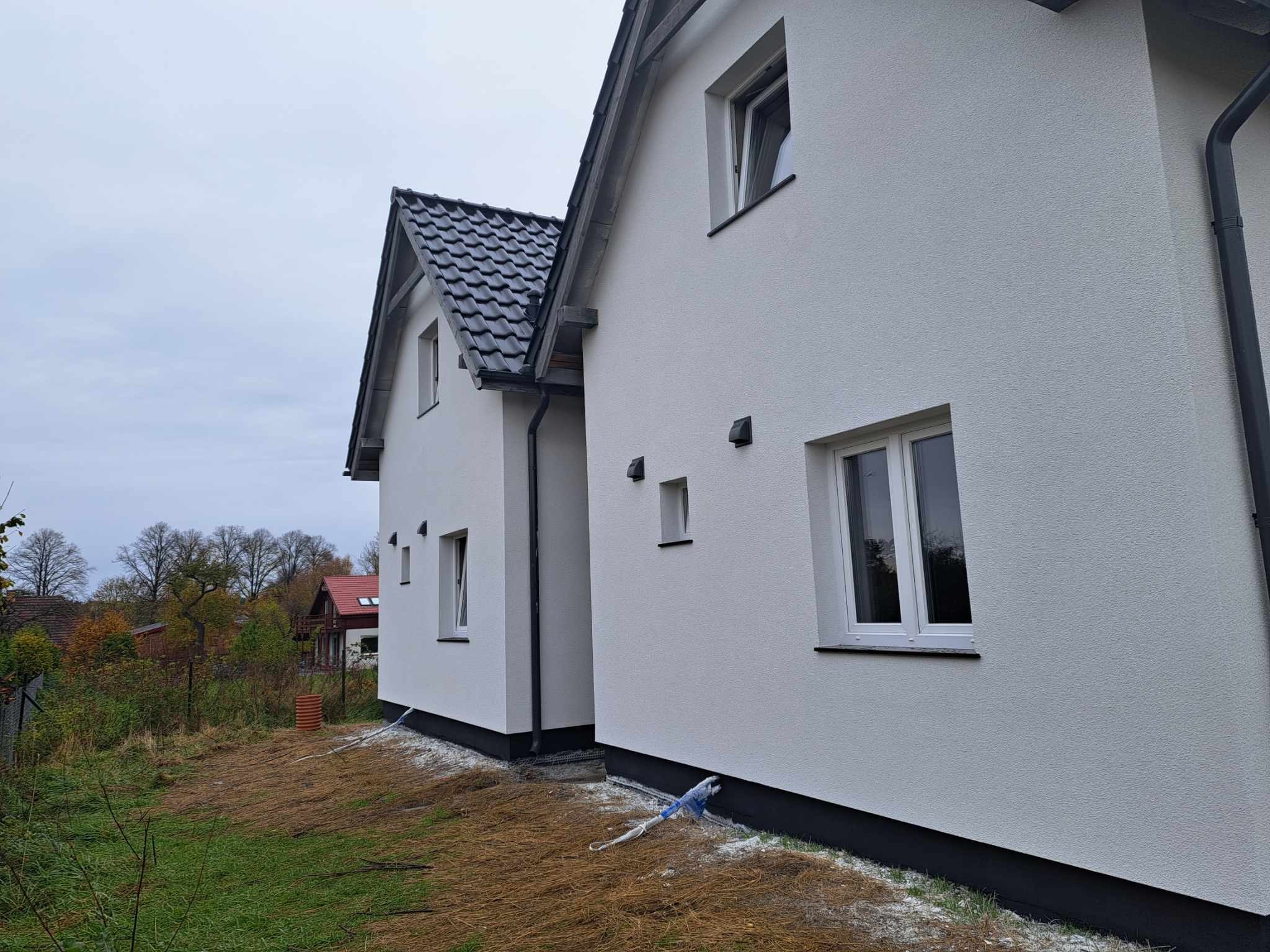 Dwa budynki jednorodzinne z białą elewacją o strukturze baranka, czarną podmurówką i grafitową dachówką, widoczne przewody i resztki styropianu po pracach.
