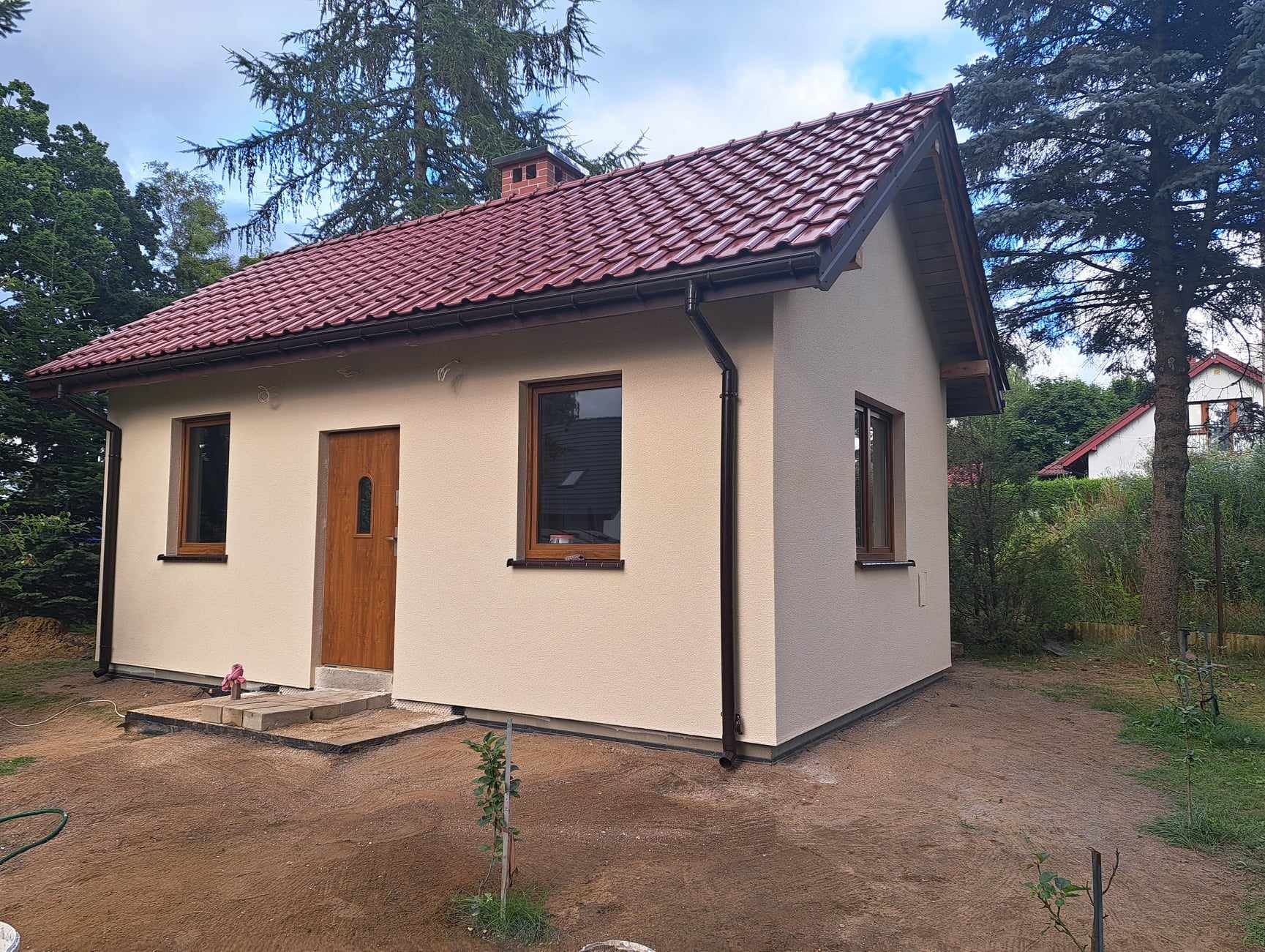 Niewielki, ocieplony dom z beżową elewacją, brązowymi oknami i drzwiami oraz czerwoną dachówką, otoczony przygotowanym terenem pod ogród.