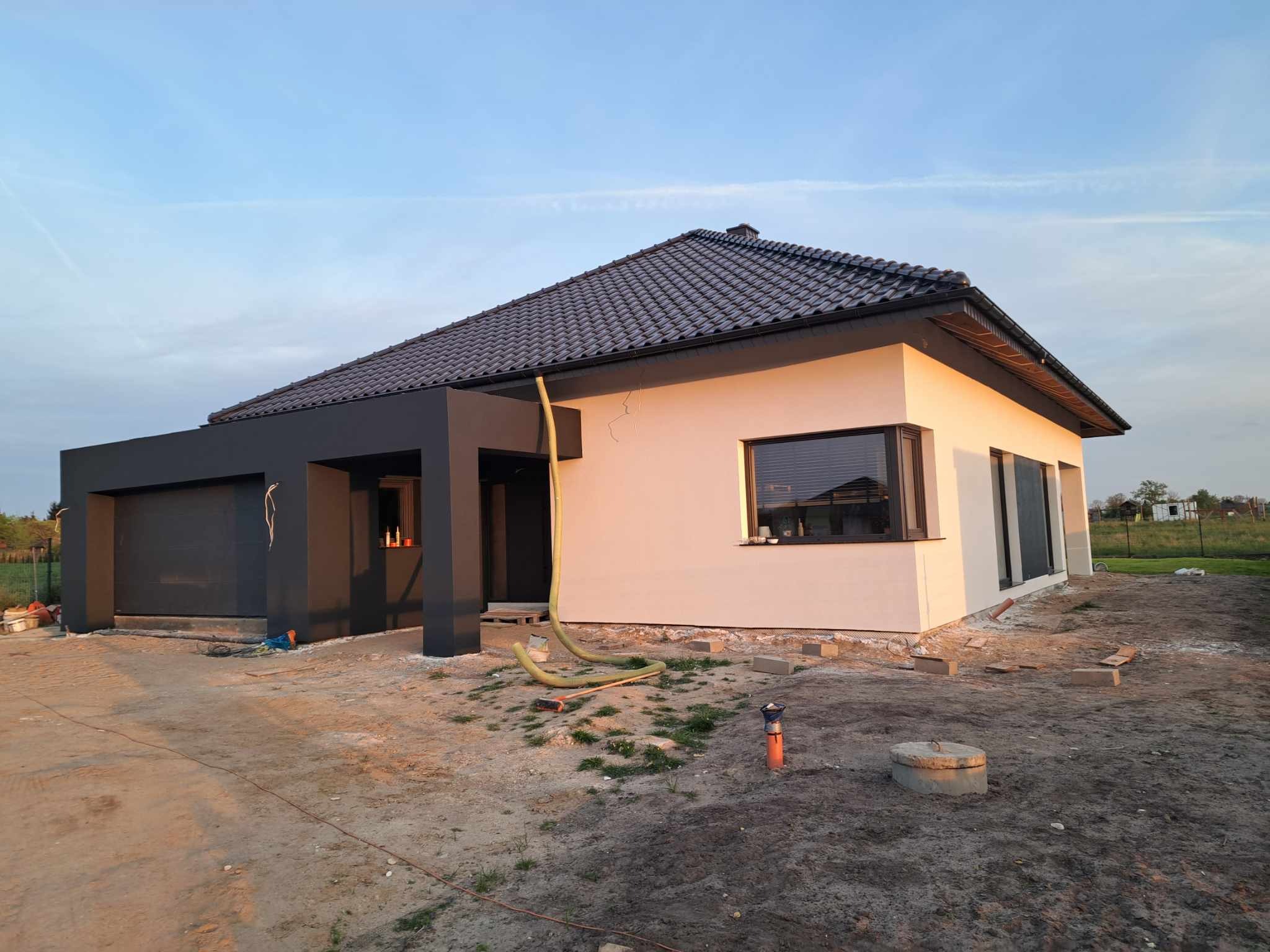 Nowoczesny dom jednorodzinny z ciemną bryłą garażu i jasną elewacją, widoczne rury wentylacyjne na zewnątrz, teren wokół domu w trakcie prac wykończeniowych.