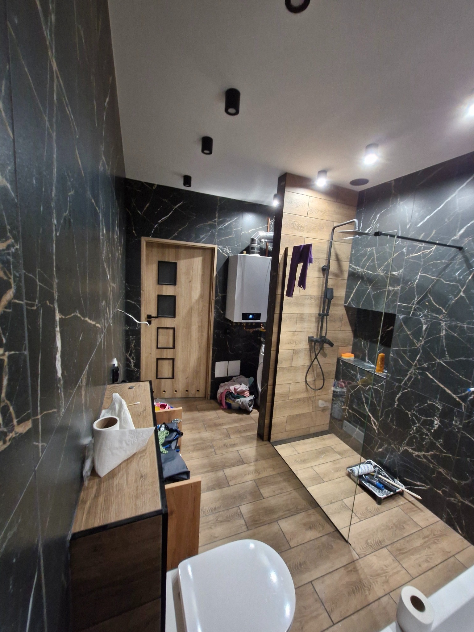 Nowoczesna łazienka z czarnymi ścianami imitującymi marmur, drewnianą podłogą i elementami wykończenia w drewnie, kabina prysznicowa ze szklaną ścianką, wiszący bojler i nieporządek na podłodze.