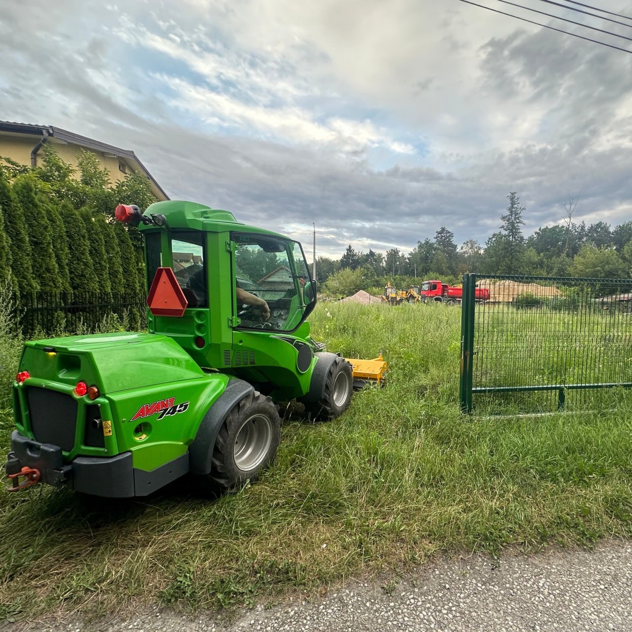 Zielony miniładowarka Avant 745 na trawiastym terenie, z widocznymi w tle wywrotką i koparką, sugerującymi prace ziemne.