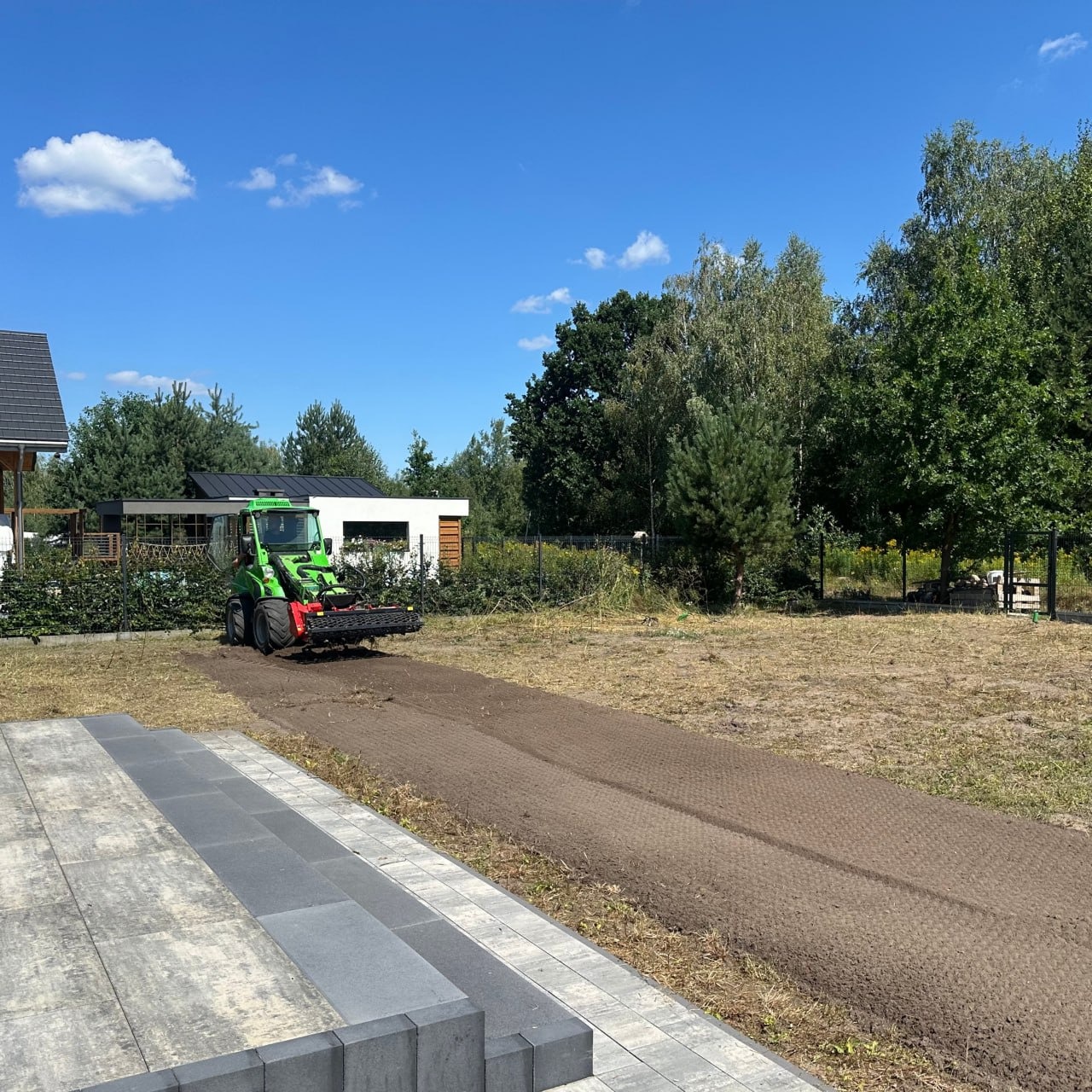 Zielony traktor z glebogryzarką przygotowuje podłoże pod trawnik na działce, widoczne obrzeże z kostki brukowej i budynek w tle.