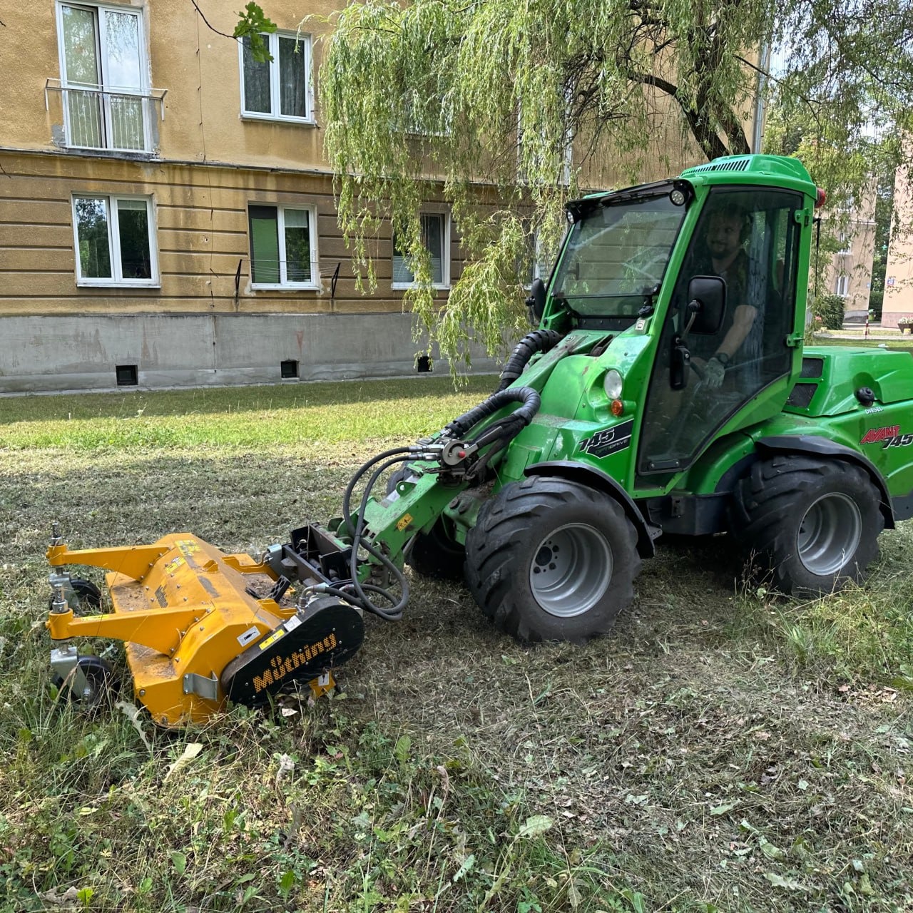 Zielony traktor z żółtą kosiarką bijakową marki Müthing na trawniku przed budynkiem mieszkalnym, widoczny operator w kabinie.
