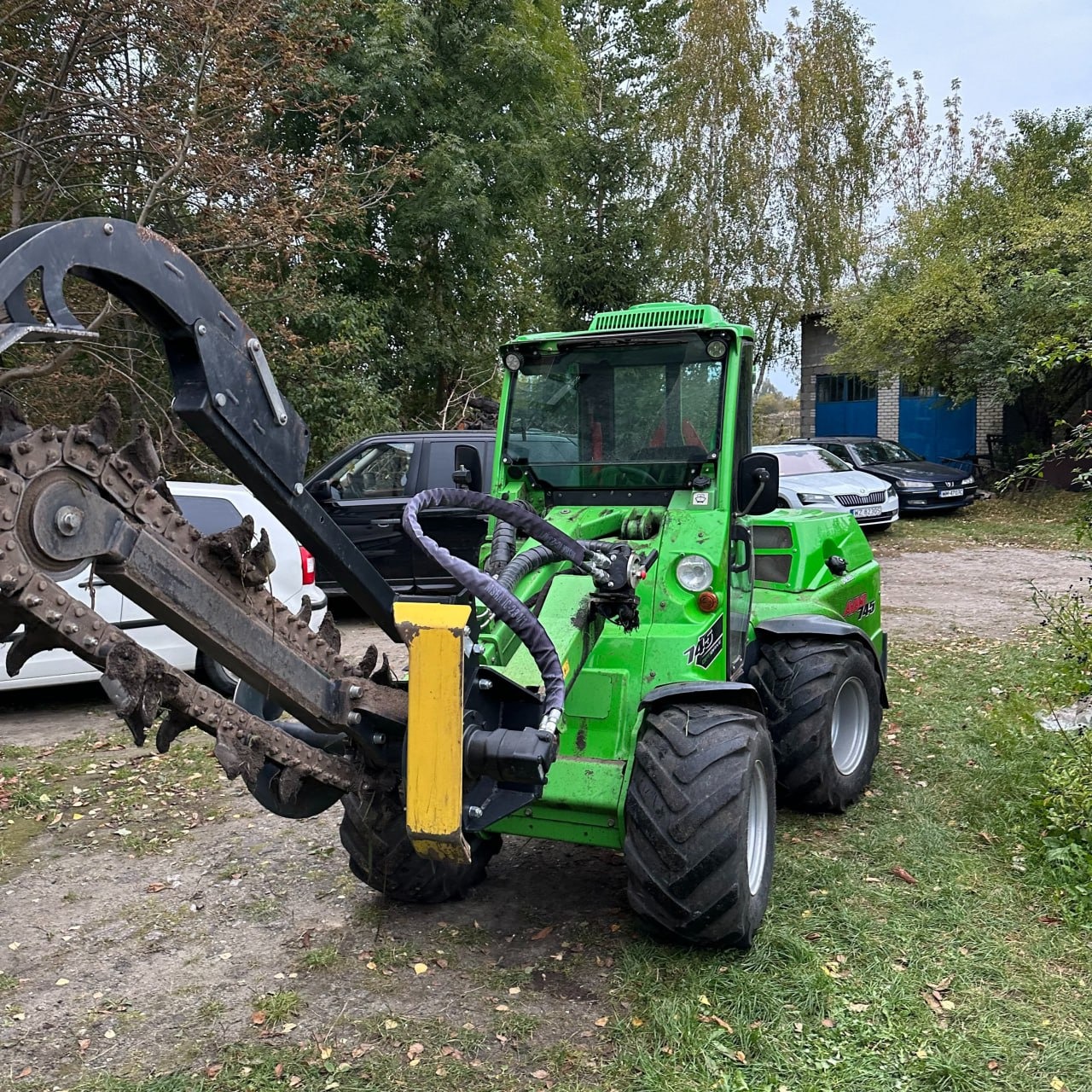 Zielony traktor z zamontowaną koparką łańcuchową stoi na trawiastym terenie, w tle widoczne budynki i samochody.