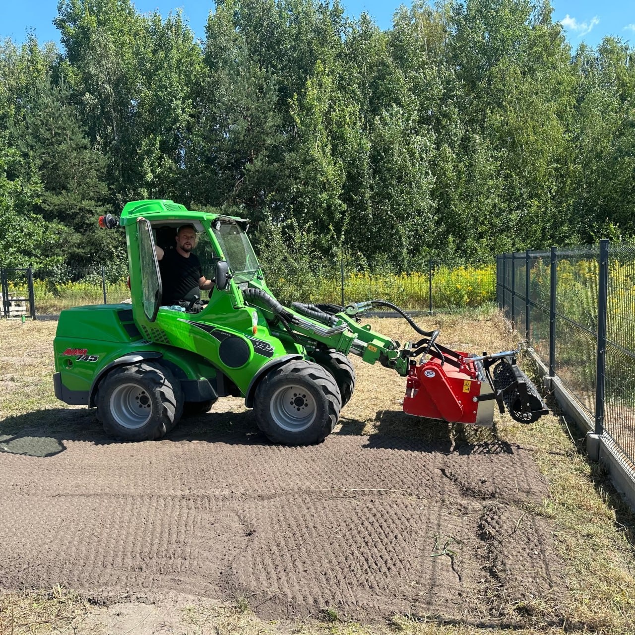 Zielona minitraktorka marki Avant 745 z czerwonym frezem glebogryzarkowym wyrównuje teren przy metalowym ogrodzeniu panelowym, przygotowując podłoże pod trawnik na tle zieleni drzew.