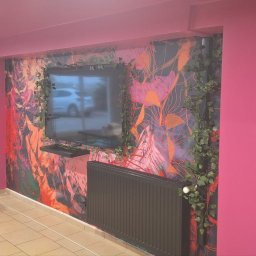 Zbigniew Utnicki - Wnętrze pokoju z pomalowanymi na różowo ścianami, dekoracyjną tapetą z motywem roślinnym za telewizorem, na tle widoczna osoba stojąca na podwyższeniu przy szklanych drzwiach.