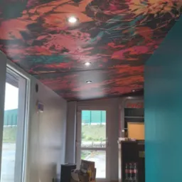 Wnętrze lokalu z pomalowanym sufitem w abstrakcyjne wzory w odcieniach czerwieni, pomarańczy i czerni, widoczne lampy sufitowe, fragment baru z lodówką Coca-Cola, jasne płytki podłogowe i turkusowa...