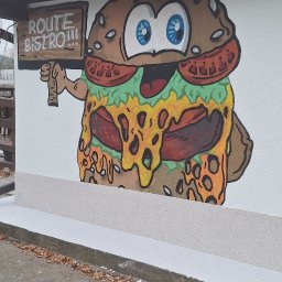 Zbigniew Utnicki - Kreskówkowy mural przedstawiający hamburgera z tabliczką 'Route Bistro!!!' na białej ścianie budynku z szarym cokołem i brązowym dachem, z widocznymi opadłymi liśćmi na ziemi.