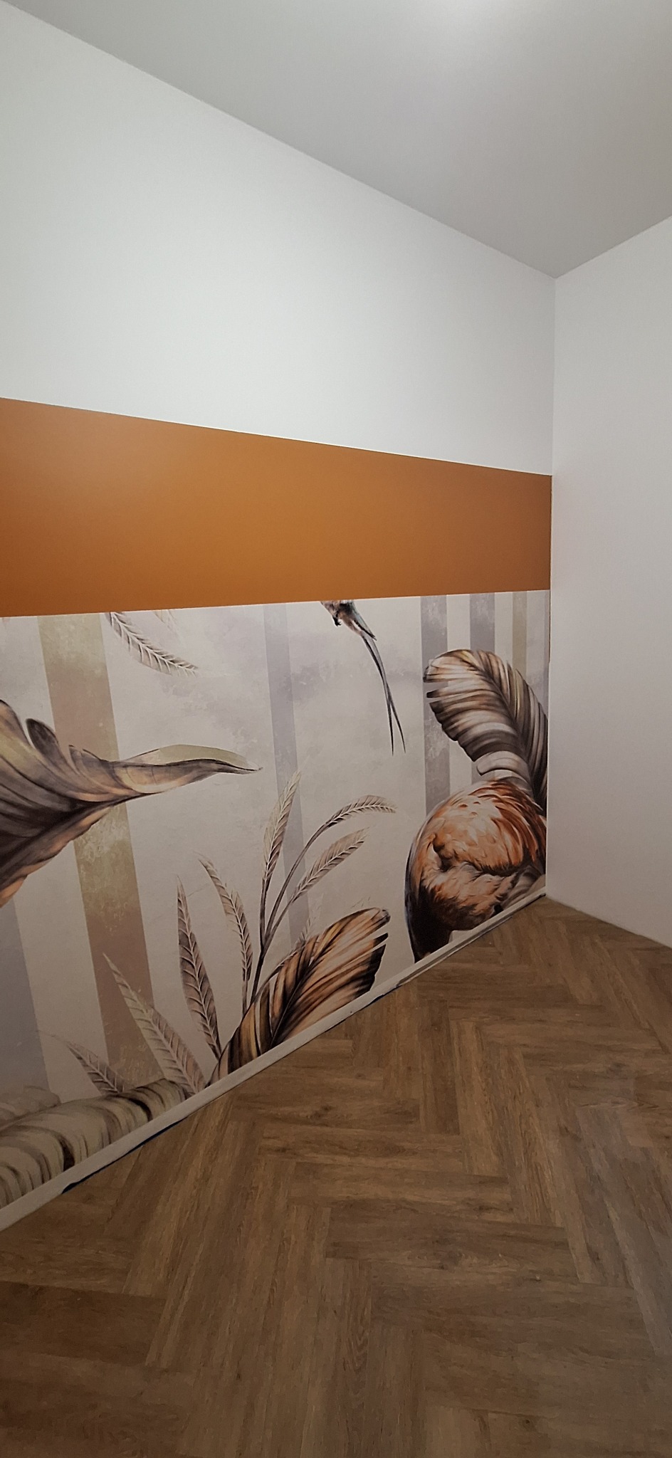 Wnętrze pokoju z panelami podłogowymi ułożonymi w jodełkę, ścianą z białym i brązowym pasem oraz tapetą w tropikalne liście i ptaki.
