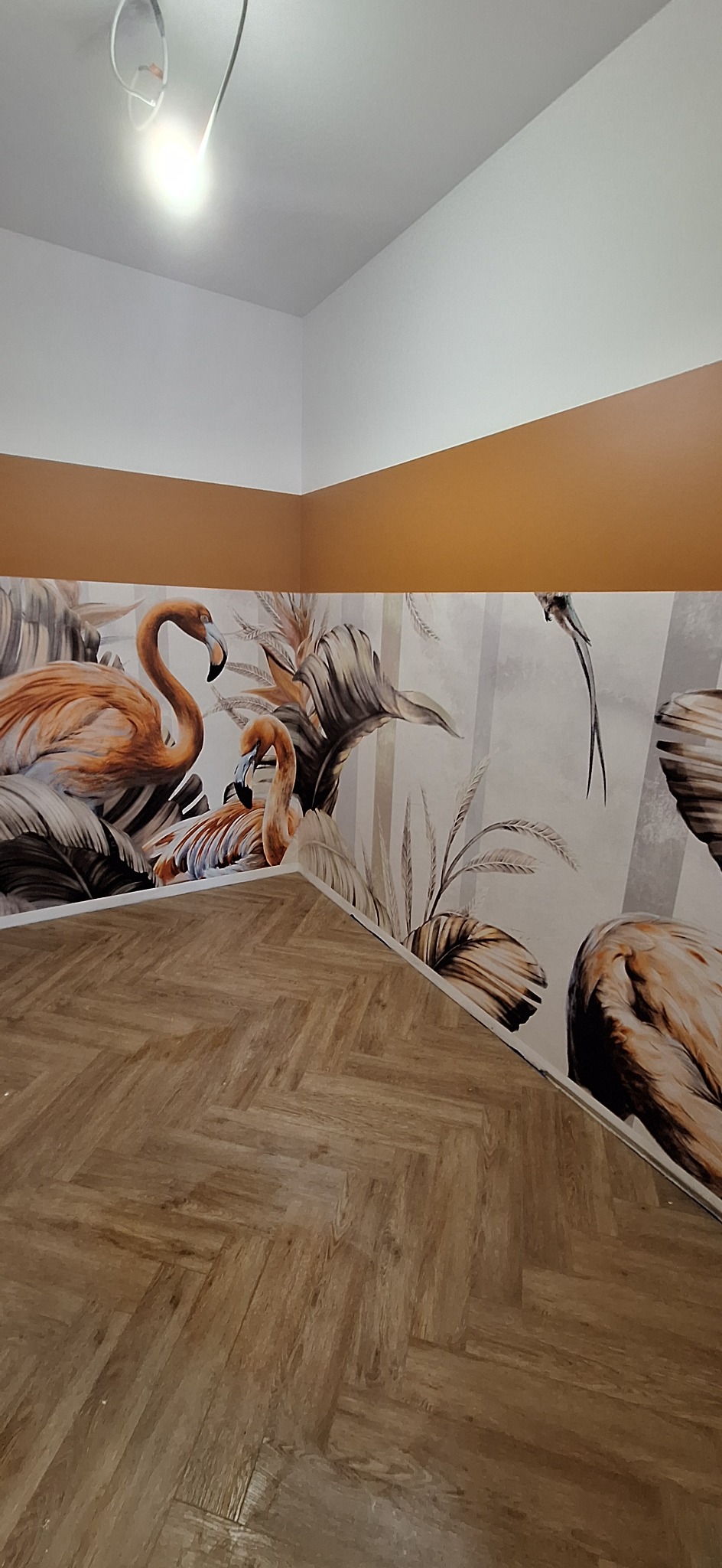 Świeżo ułożona podłoga z paneli w jodełkę sąsiaduje z pomalowaną ścianą z motywem flamingów i roślin tropikalnych, oddzieloną od białego sufitu i brązowej ściany pasem w kolorze karmelowym.