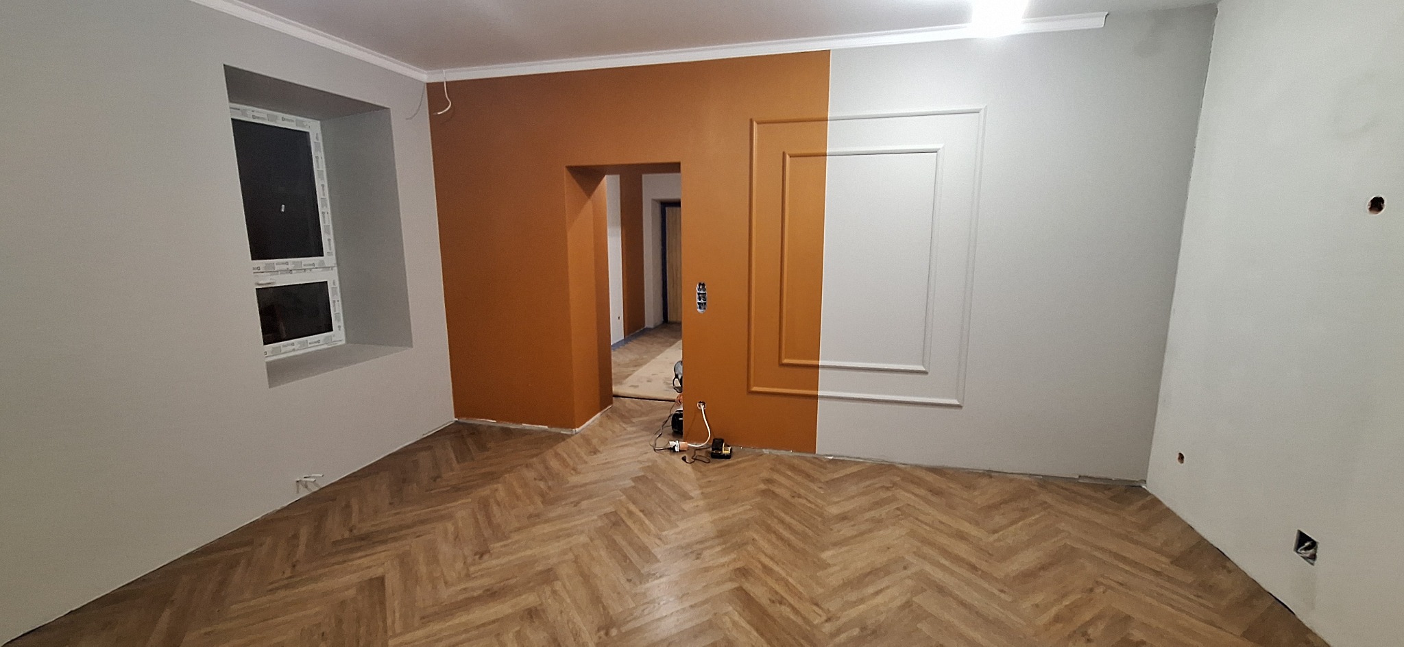 Pokój po malowaniu z panelami ułożonymi w jodełkę, ściany pomalowane na szaro i pomarańczowo, dekoracyjne listwy ścienne, widoczne gniazdka elektryczne i sprzęt malarski.