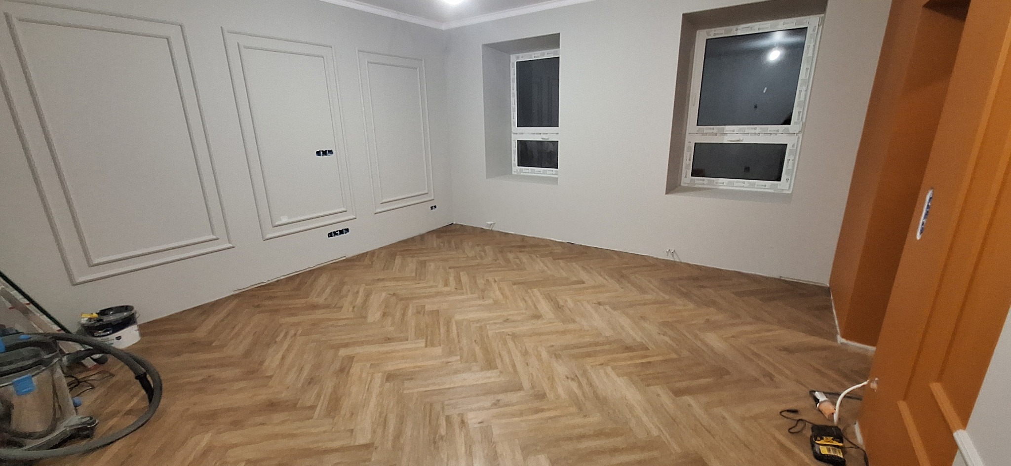 Pokój po remoncie z ułożonymi panelami w jodełkę, ściany pomalowane na szaro z dekoracyjnymi listwami, widoczne dwa okna i częściowo drzwi w kolorze pomarańczowym, na podłodze leży odkurzacz...