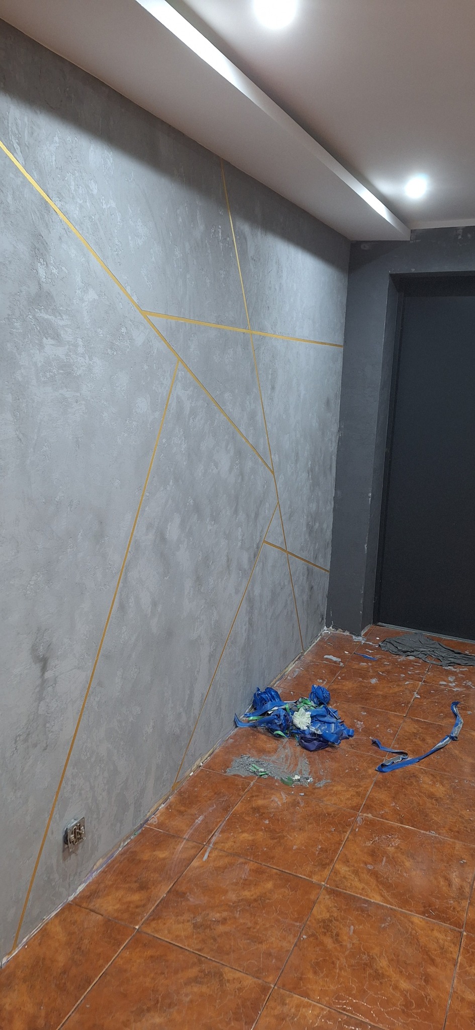 Szara ściana z dekoracyjnymi złotymi liniami, brązowa podłoga z resztkami farby i niebieską folią ochronną, czarne drzwi w tle.