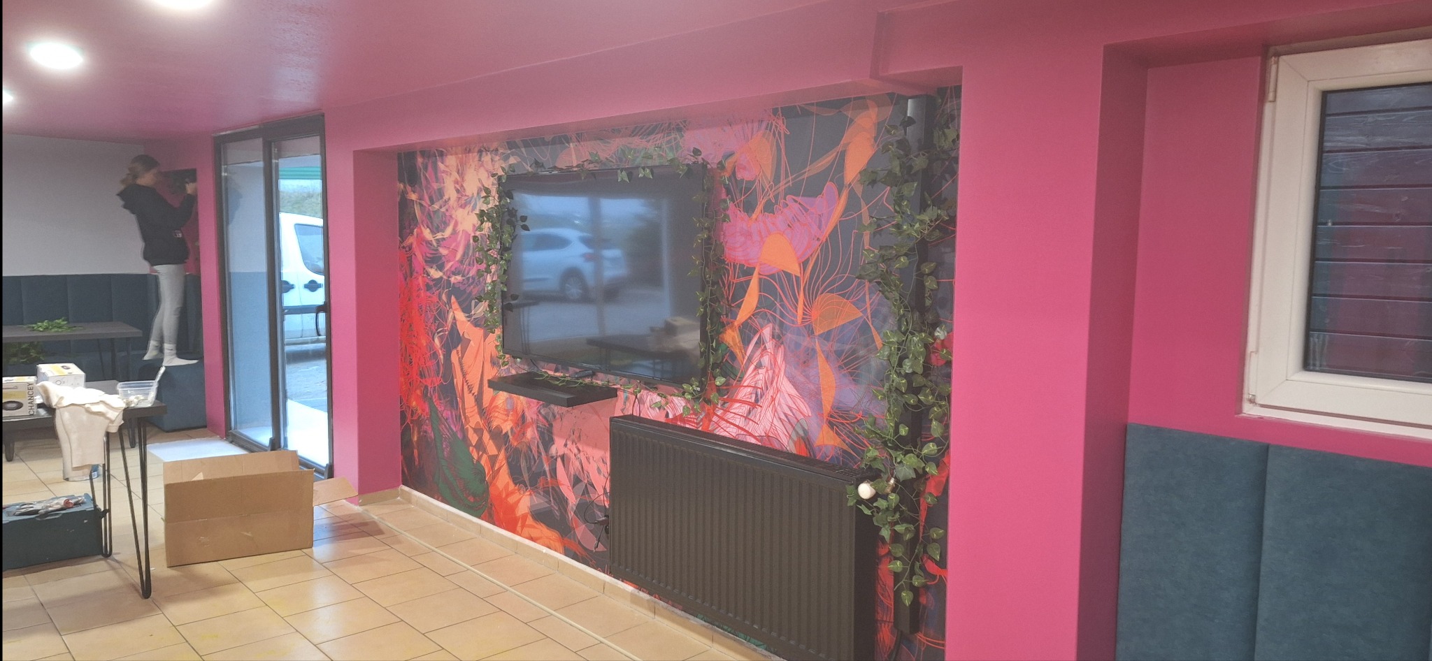 Wnętrze pokoju z pomalowanymi na różowo ścianami, dekoracyjną tapetą z motywem roślinnym za telewizorem, na tle widoczna osoba stojąca na podwyższeniu przy szklanych drzwiach.