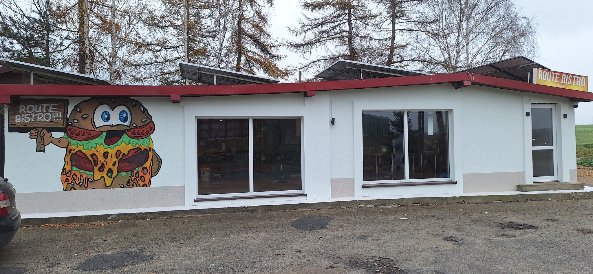 Biały budynek z czerwonym dachem, na ścianie mural przedstawiający burgera z napisem Route Bistro, okna z białymi ramami, panele słoneczne na dachu.