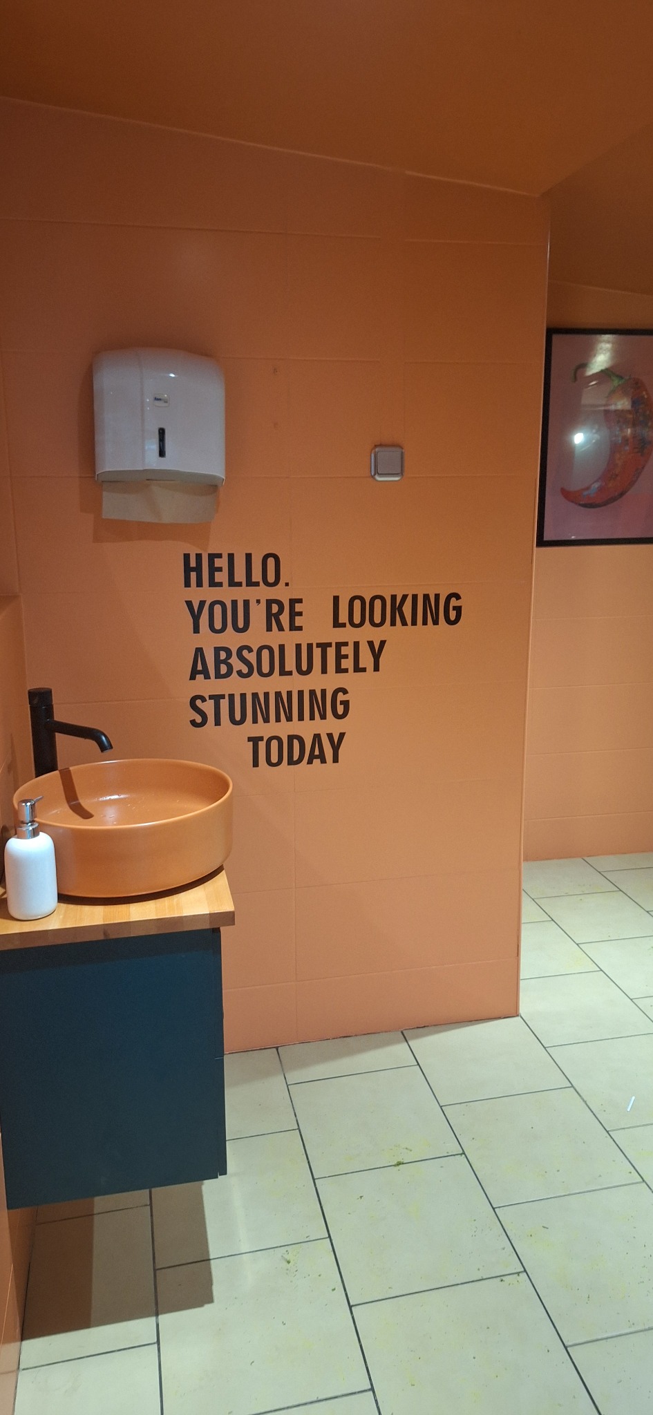 Pomarańczowa łazienka z okrągłą umywalką i czarną armaturą, na ścianie napis 'Hello. You're looking absolutely stunning today', dozownik na papier i obrazek z papryczką chili.