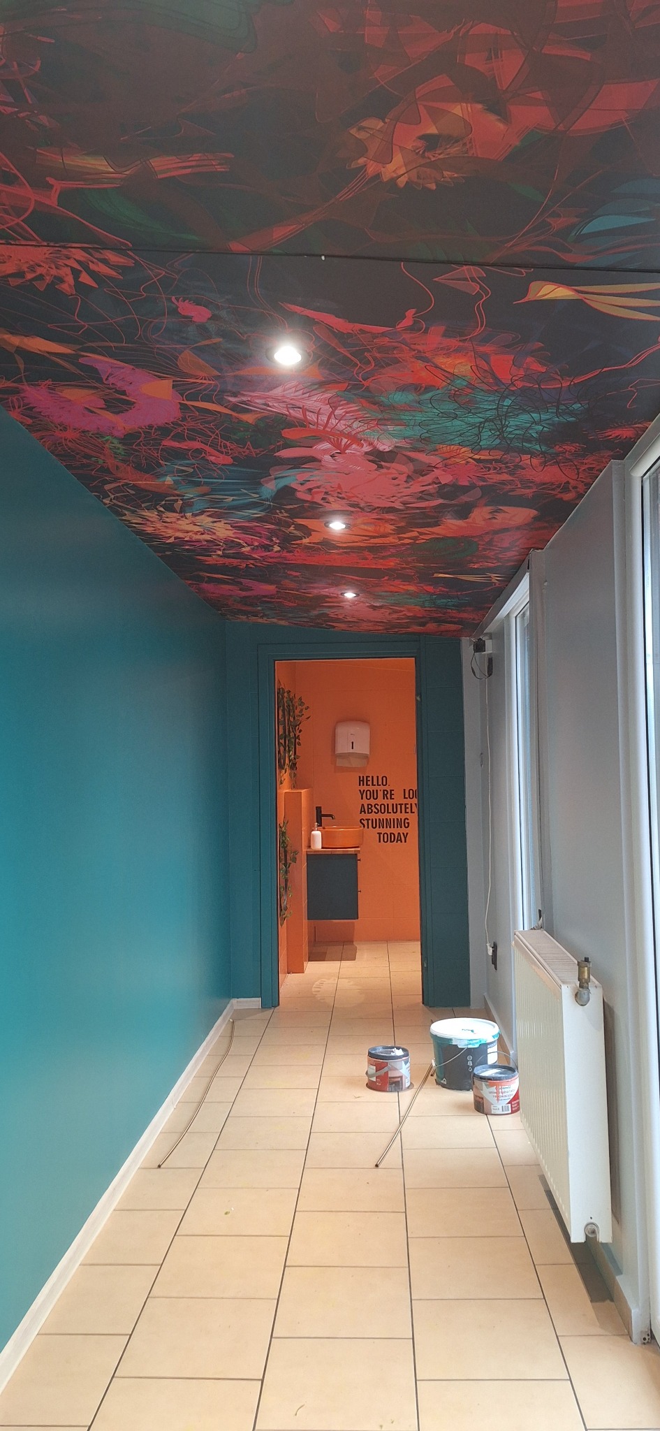 Wnętrze biura z pomalowanymi ścianami na turkusowo i sufitem z abstrakcyjnym wzorem, widoczne otwarte drzwi do pomalowanej na pomarańczowo łazienki z napisem motywacyjnym oraz puszki z farbą...