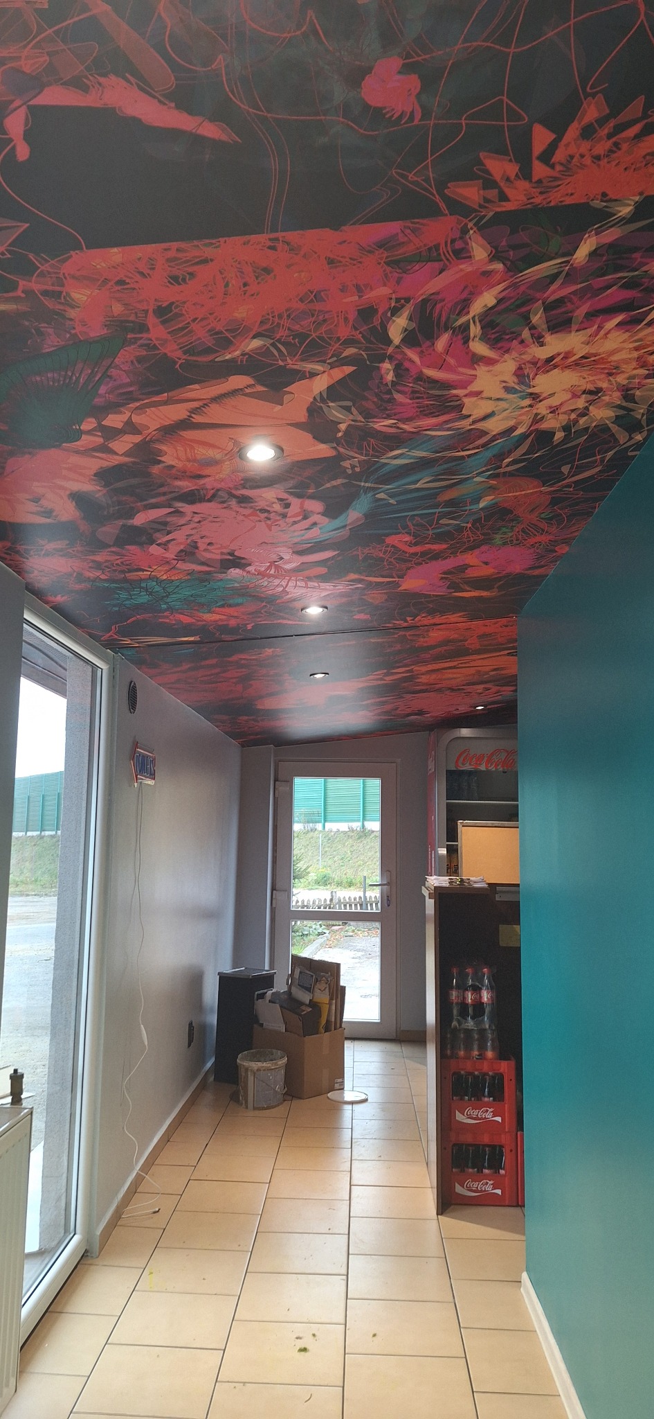 Wnętrze lokalu z pomalowanym sufitem w abstrakcyjne wzory w odcieniach czerwieni, pomarańczy i czerni, widoczne lampy sufitowe, fragment baru z lodówką Coca-Cola, jasne płytki podłogowe i turkusowa...