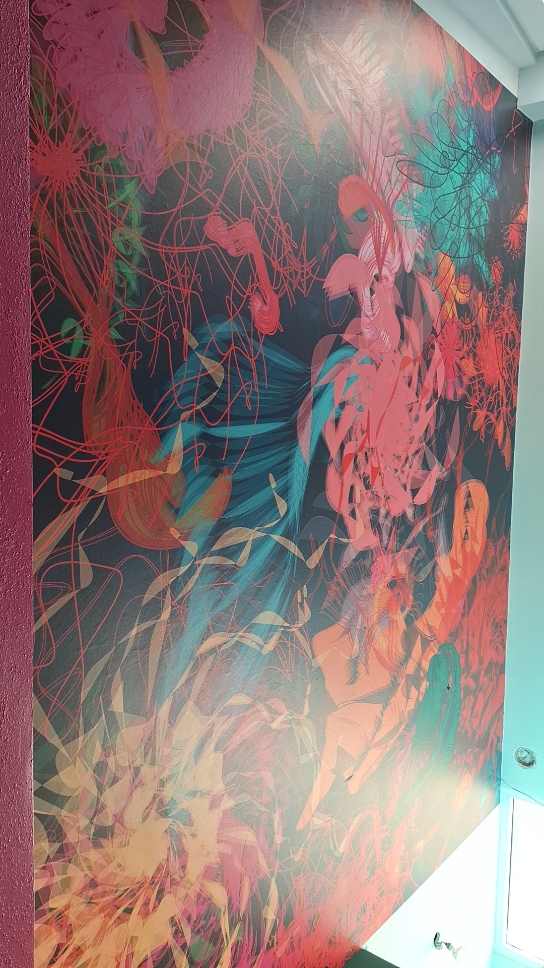 Ściana z abstrakcyjnym, kolorowym malowidłem w odcieniach różu, czerwieni, turkusu i beżu na ciemnym tle, z widocznym fragmentem fioletowej ściany bocznej i białego sufitu.