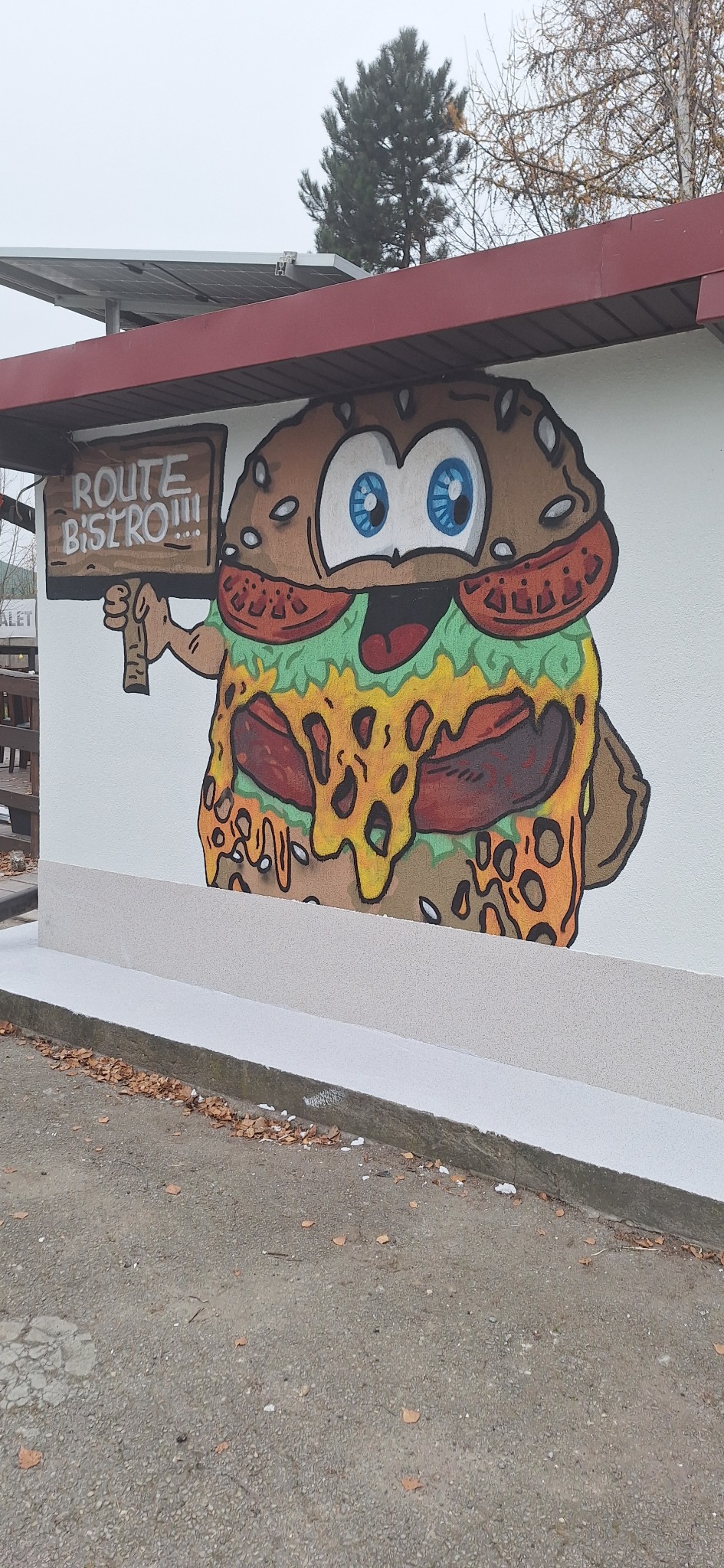 Kreskówkowy mural przedstawiający hamburgera z tabliczką 'Route Bistro!!!' na białej ścianie budynku z szarym cokołem i brązowym dachem, z widocznymi opadłymi liśćmi na ziemi.