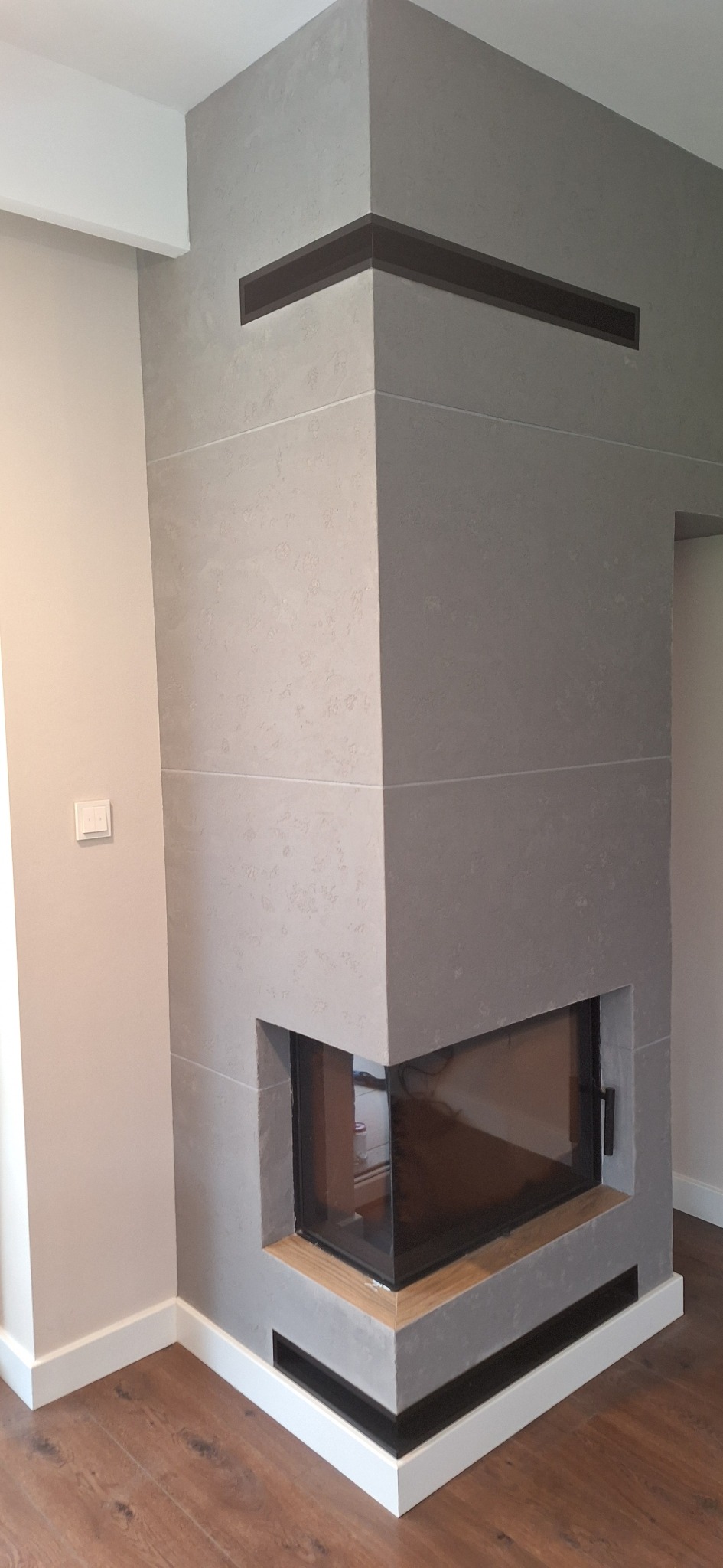 Szary, narożny kominek z wkładem kominkowym, obudowany płytami imitującymi beton, z czarnymi wlotami powietrza u góry i dołu, na tle ściany pomalowanej na kremowo z białymi listwami przypodłogowymi...