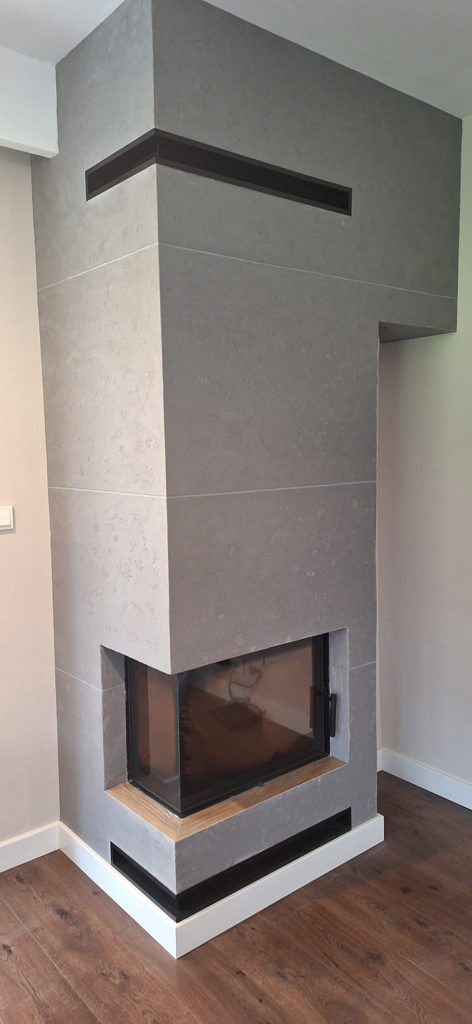 Nowoczesny kominek narożny z szarą, teksturowaną obudową, drewnianymi akcentami i czarnymi wstawkami wentylacyjnymi, na tle parkietu.