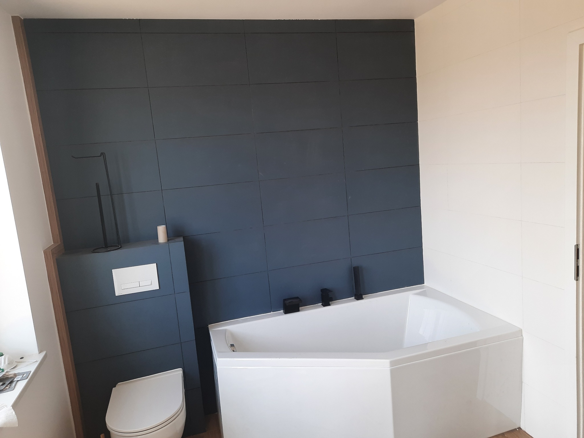 Nowoczesna łazienka z ciemnymi, kwadratowymi płytkami na ścianie, białą wanną narożną i podwieszaną toaletą. Bateria wannowa i akcesoria w kolorze czarnym.