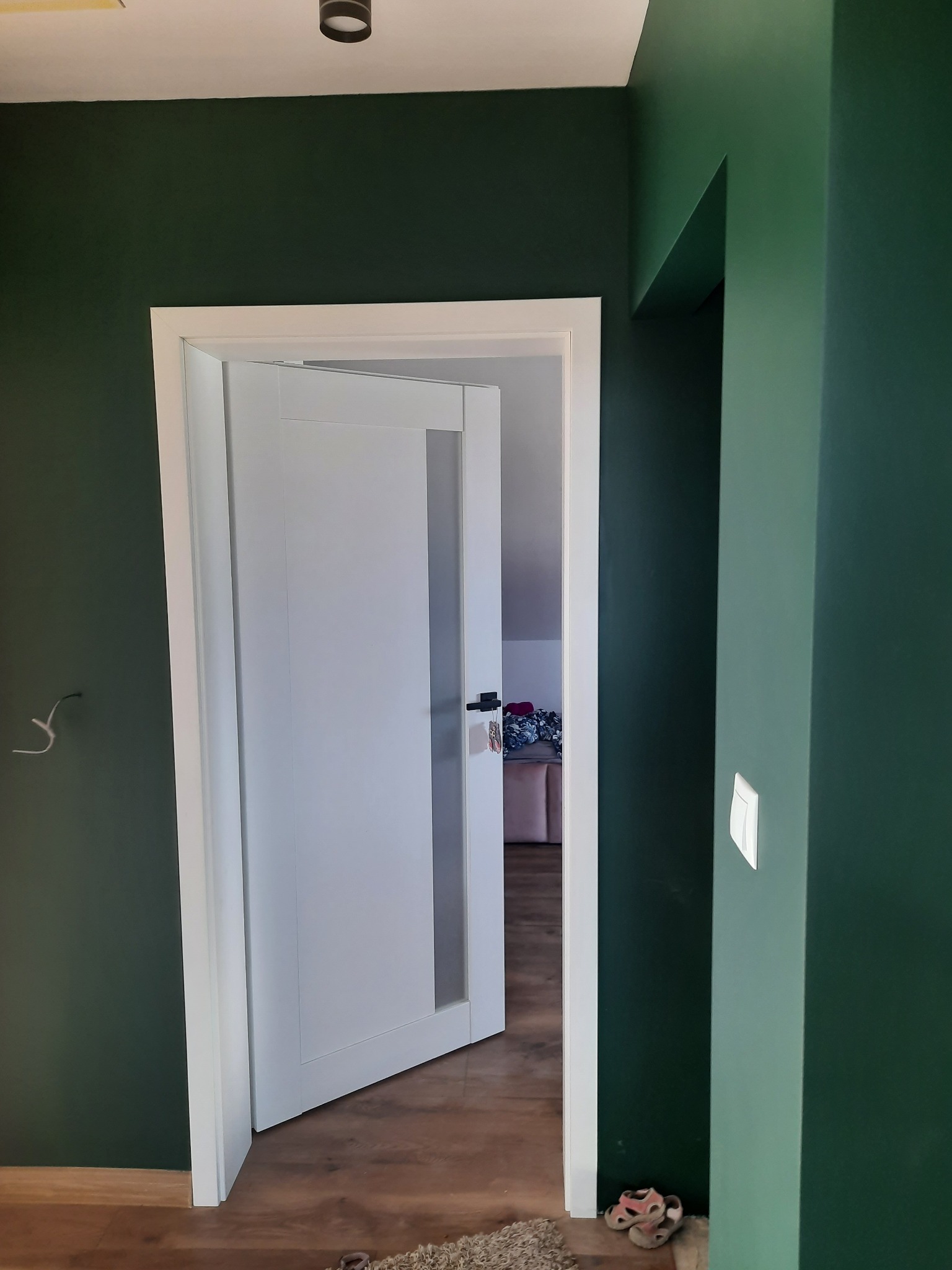 Otwarta, biała, wewnętrzna stolarka drzwiowa z przeszkleniem w zielonym pokoju, widoczny fragment podłogi i przełącznik światła.
