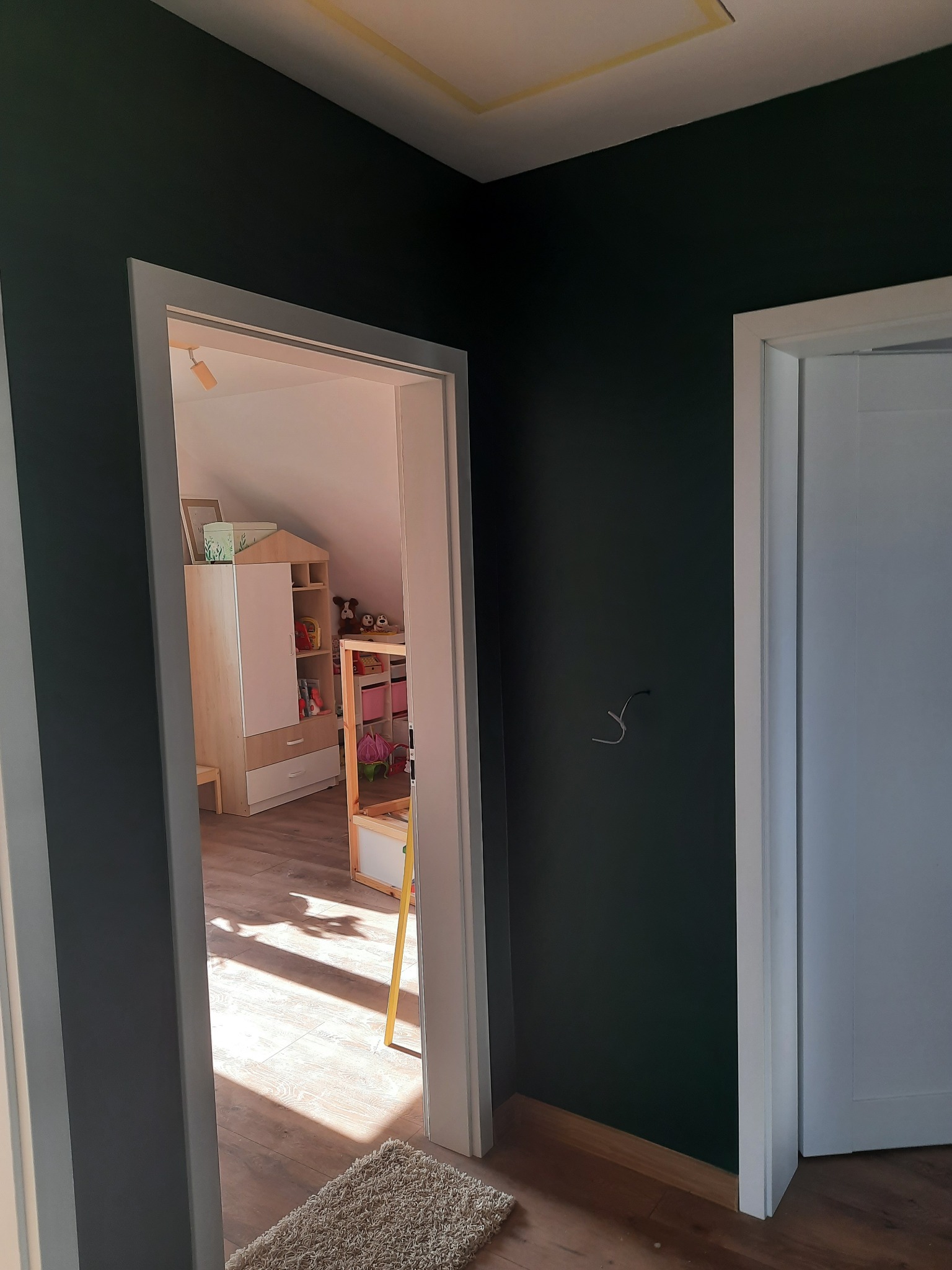 Ciemnozielona ściana w pokoju dziecięcym z widocznym fragmentem białych drzwi i wejściem do pokoju z drewnianą podłogą, meblami i zabawkami, oświetlonym promieniami słońca.