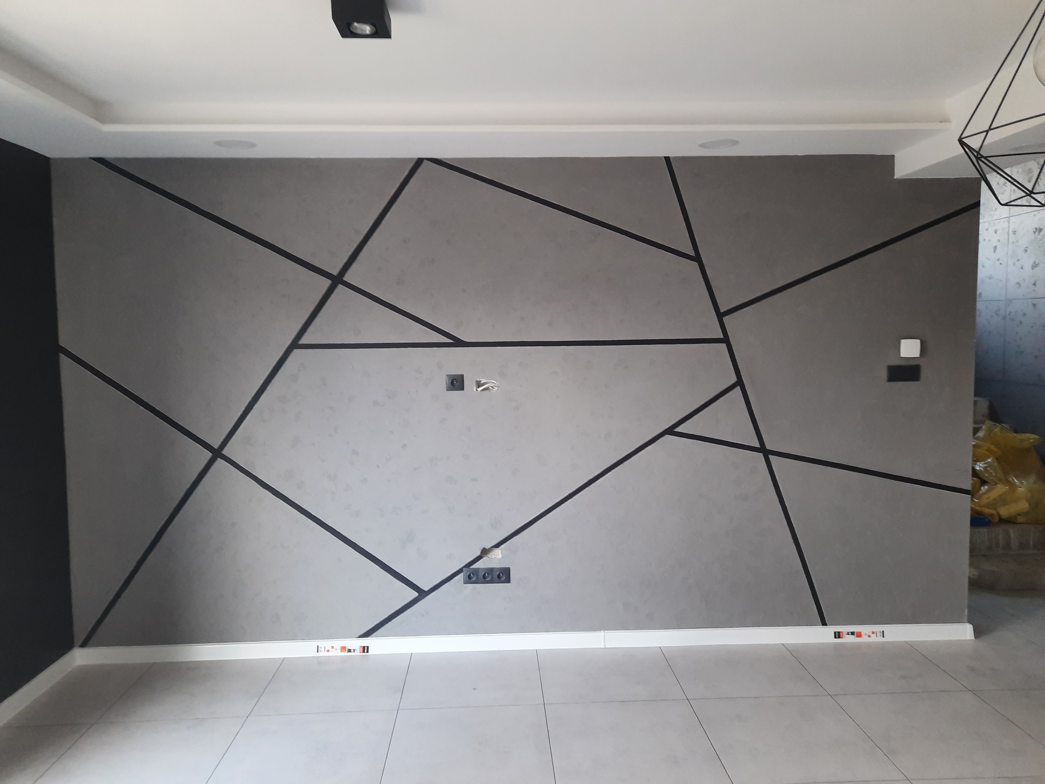 Szara ściana z dekoracyjnymi czarnymi liniami tworzącymi geometryczny wzór, widoczne gniazdka elektryczne i poziomice przy listwach przypodłogowych.