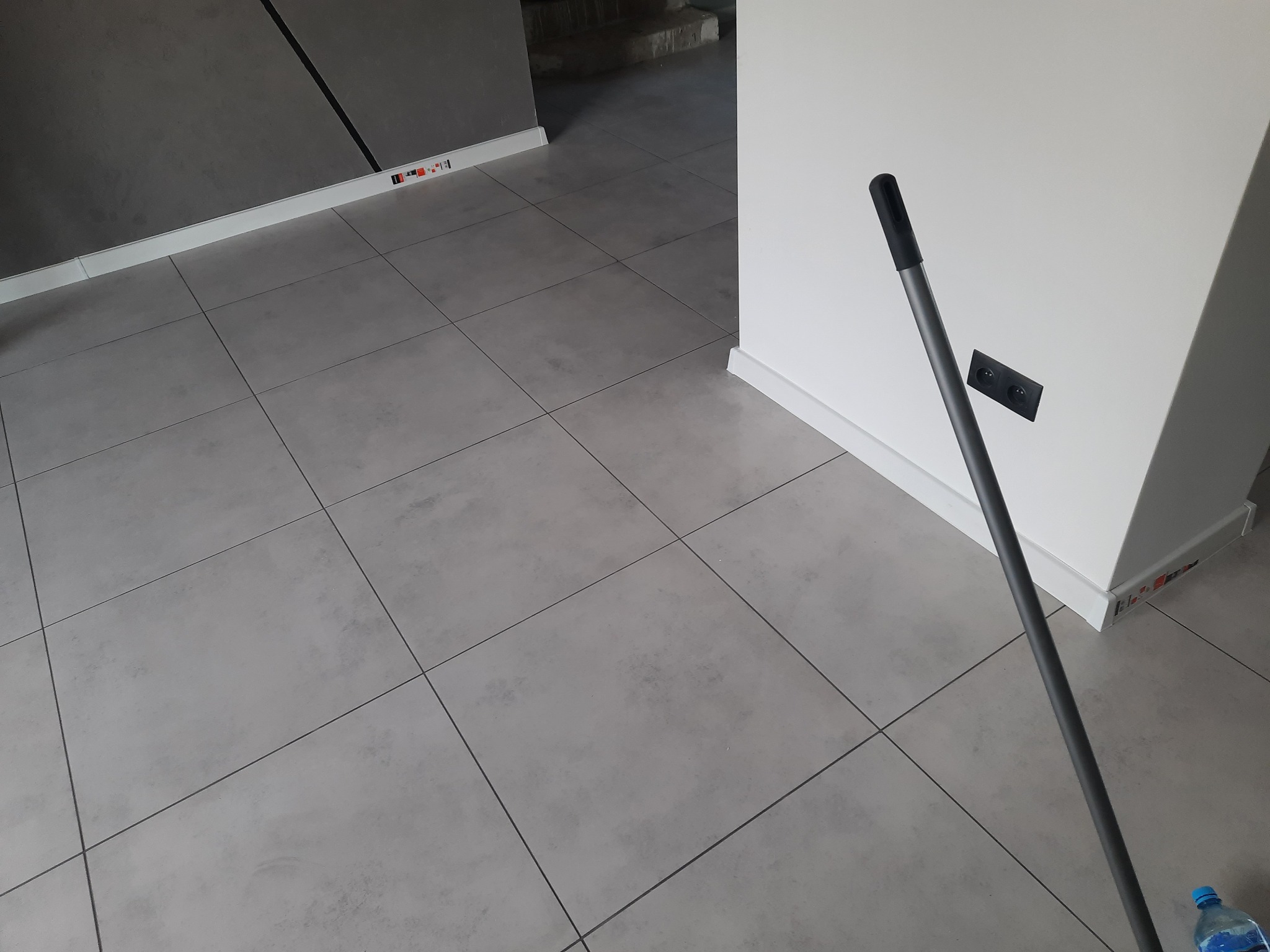 Szare, matowe płytki podłogowe ułożone w regularną siatkę, widoczne listwy przypodłogowe i fragment ściany z gniazdkiem elektrycznym oraz mop oparty o ścianę