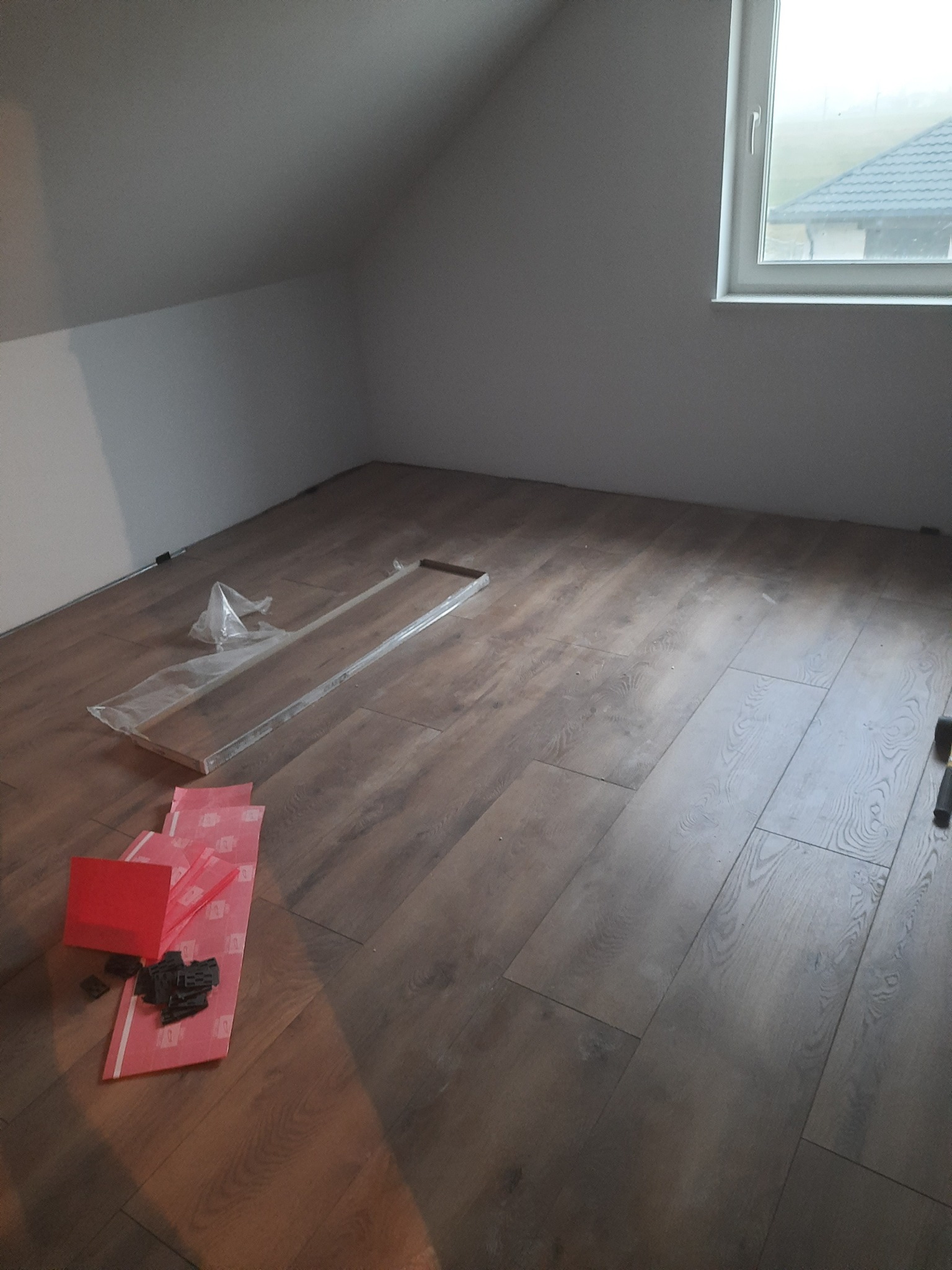 Fragment poddasza z częściowo ułożonymi panelami podłogowymi w odcieniach brązu, widoczne akcesoria montażowe, folia i narzędzia, w tle okno dachowe.