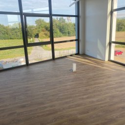 KATRUK Technologies - Nowa, pusta przestrzeń biurowa z panelami podłogowymi w odcieniach brązu, widok na zieleń przez duże okna, minimalistyczne oświetlenie sufitowe i białe ściany.