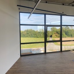 KATRUK Technologies - Nowoczesne wnętrze biurowe z dużą ścianą okienną wychodzącą na zielony krajobraz, widoczna podłoga imitująca drewno i surowy betonowy sufit z oświetleniem liniowym.