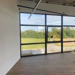 Nowoczesne wnętrze biurowe z dużą ścianą okienną wychodzącą na zielony krajobraz, widoczna podłoga imitująca drewno i surowy betonowy sufit z oświetleniem liniowym.