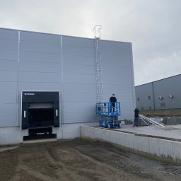 KATRUK Technologies - Montaż drabiny pionowej na elewacji hali magazynowej z wykorzystaniem podnośnika koszowego, widoczny pracownik w czarnej kurtce, drugi pracownik na ziemi, elementy elewacji w kolorze szarym.