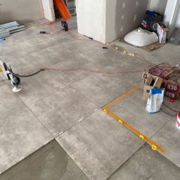 KATRUK Technologies - Układanie szarych płytek podłogowych w nowym pomieszczeniu z użyciem poziomicy laserowej i systemu poziomowania płytek, widoczne worki z zaprawą i niezamontowana umywalka.