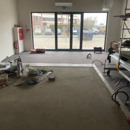 KATRUK Technologies - Przygotowanie podłoża pod posadzkę przemysłową we Wrocławiu: widok na surową wylewkę, worki z zaprawą, drabina malarska i okno z widokiem na ulicę.